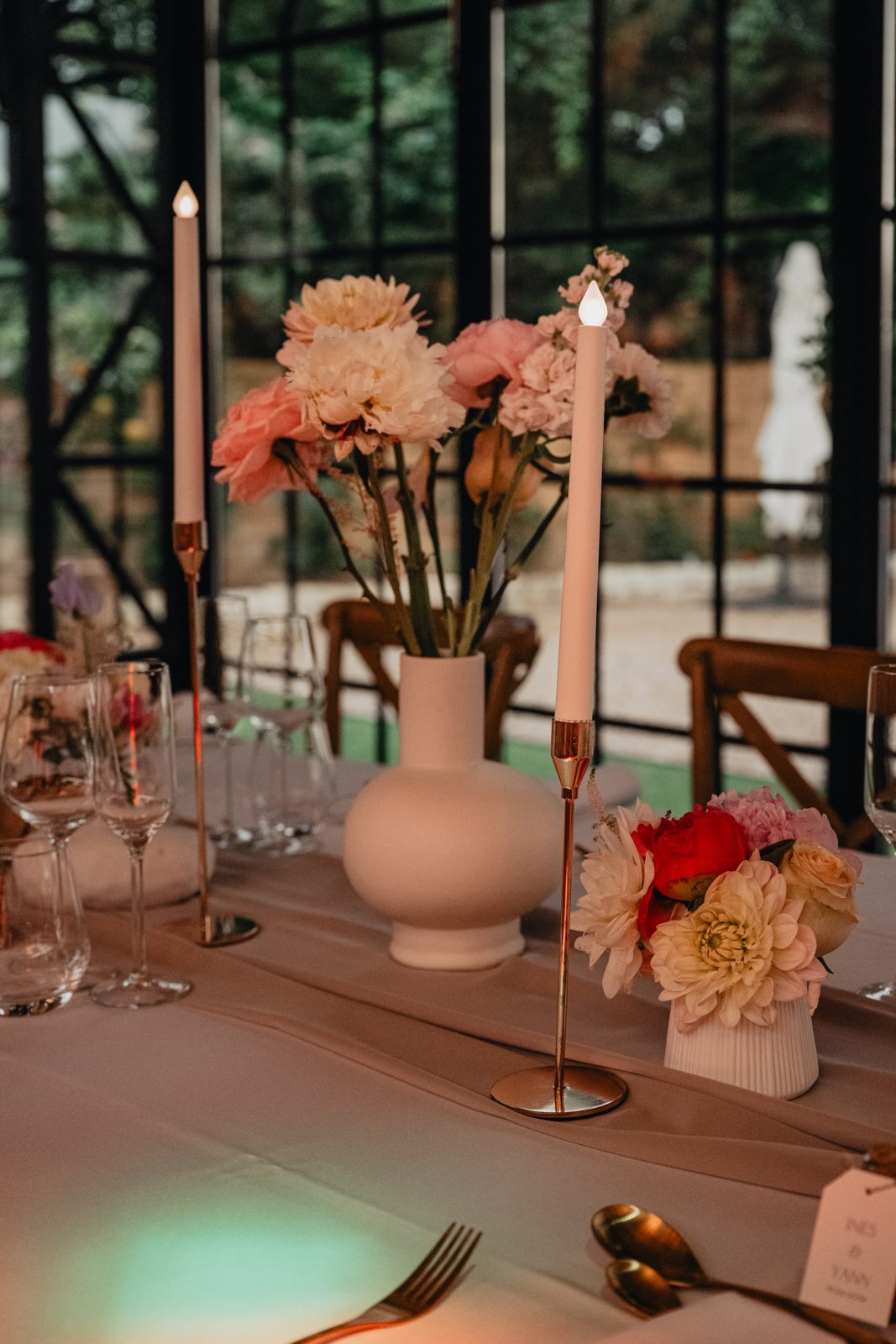 Centre de table : vase blanc de fleurs pastel, petit vase cannelé de roses et bougies sur bougeoirs dorés sur pied