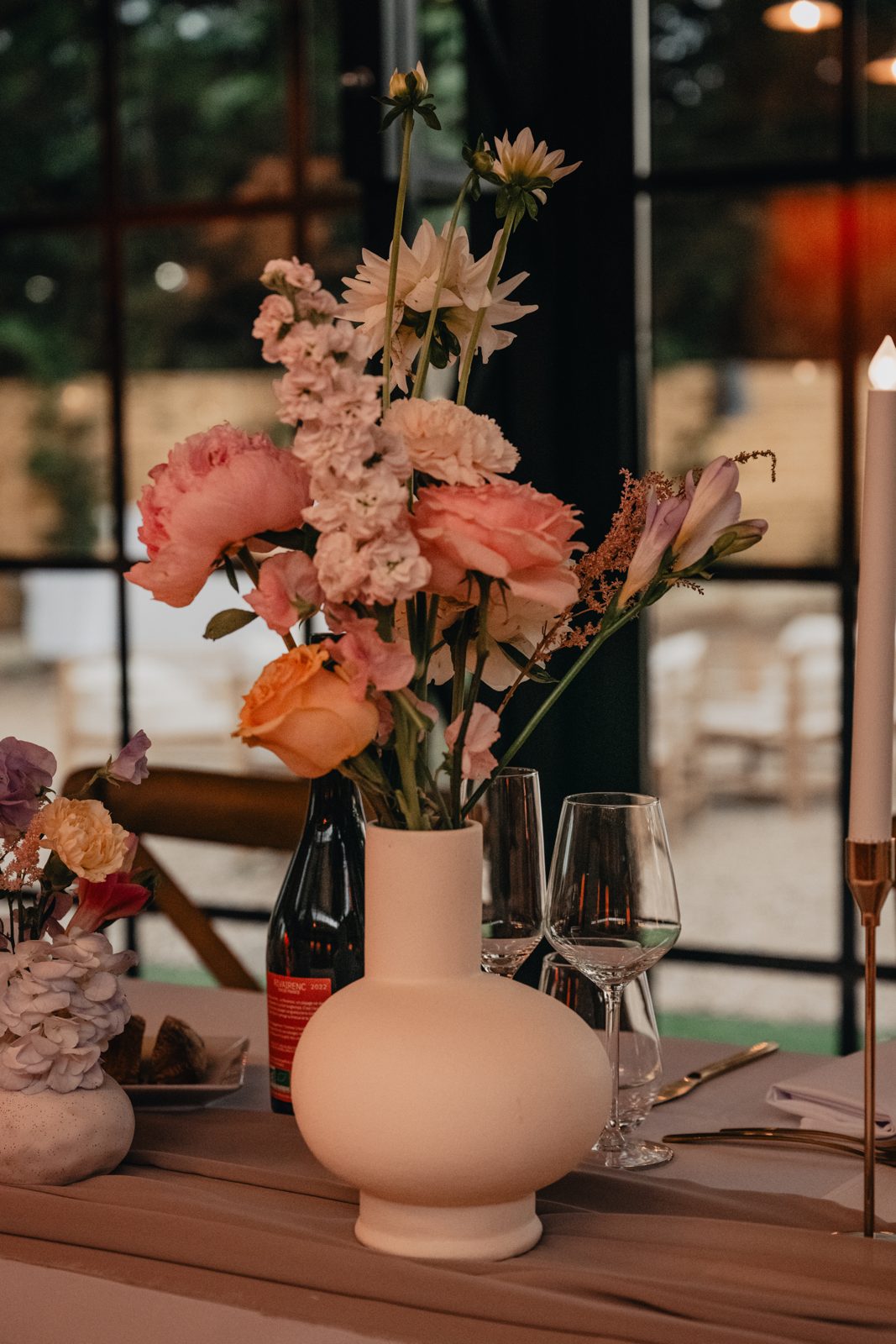 Vase blanc arrondi garni de roses, œillets et fleurs des champs sur une table dressée, bougie sur bougeoir doré