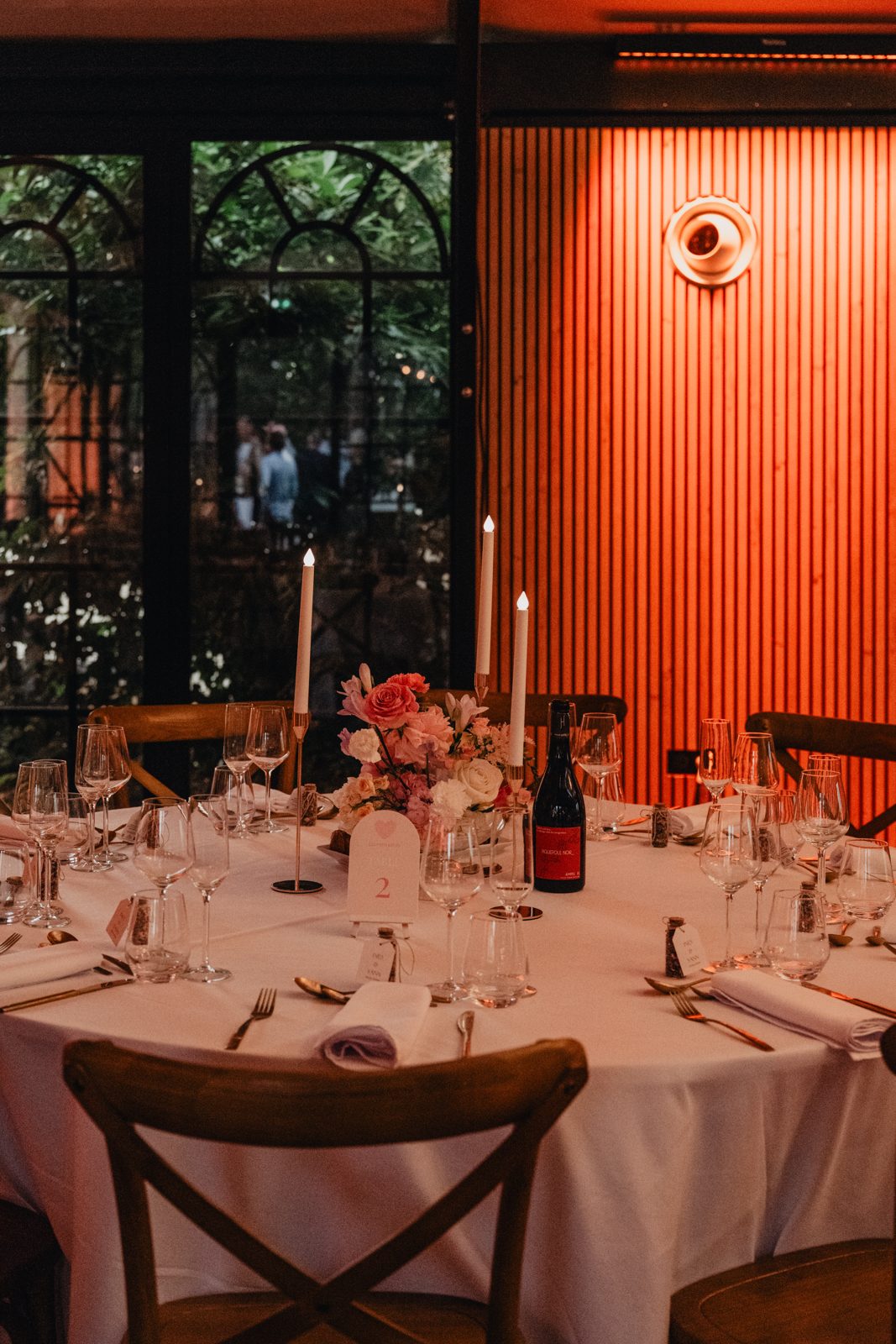 Table ronde nappée de blanc, chaises bistrot en bois, bouquet de roses et bougies sur bougeoirs dorés, mur éclairé en orange