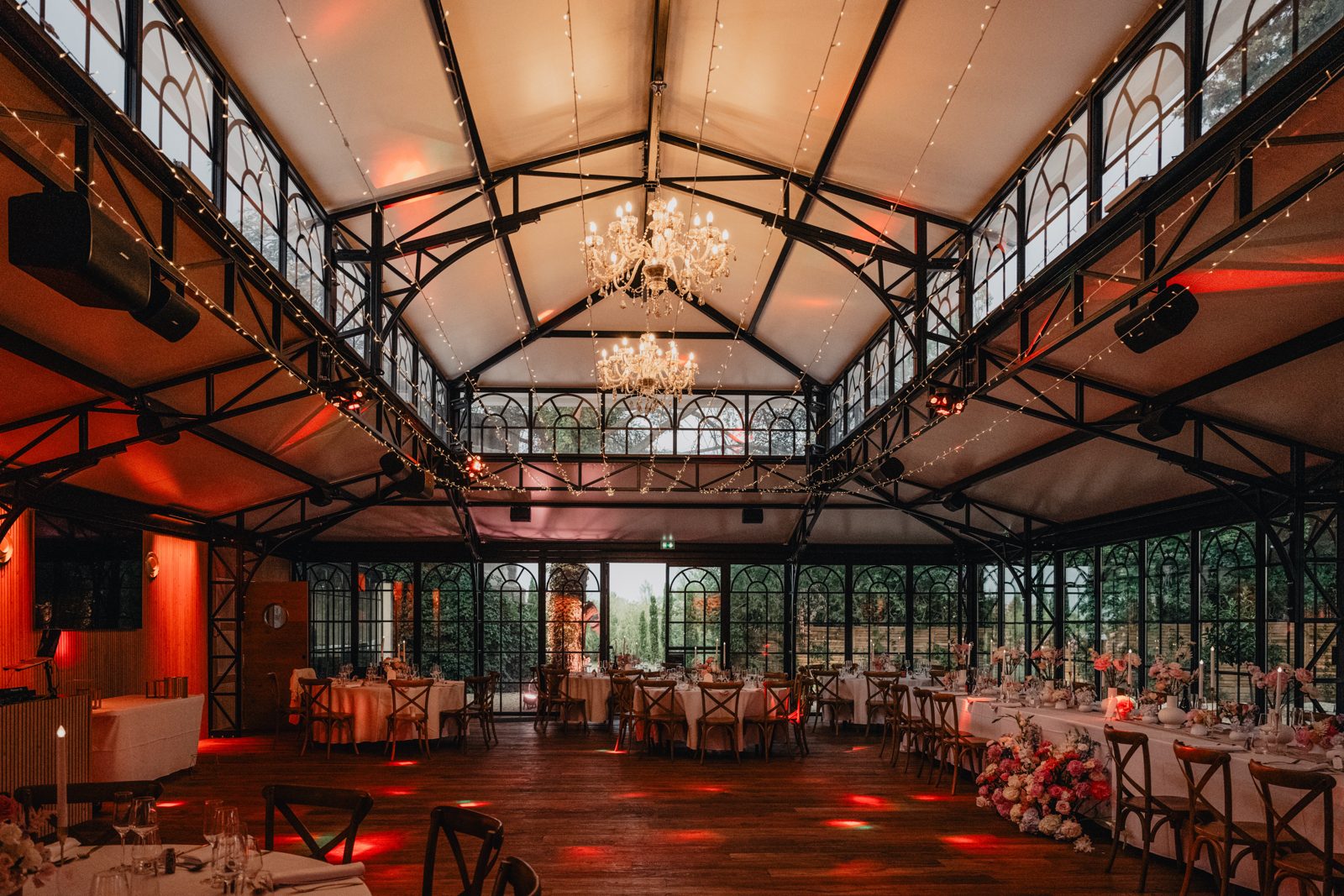 Salle de réception sous verrière avec lustres, guirlandes lumineuses, tables rondes nappées de blanc et chaises en bois