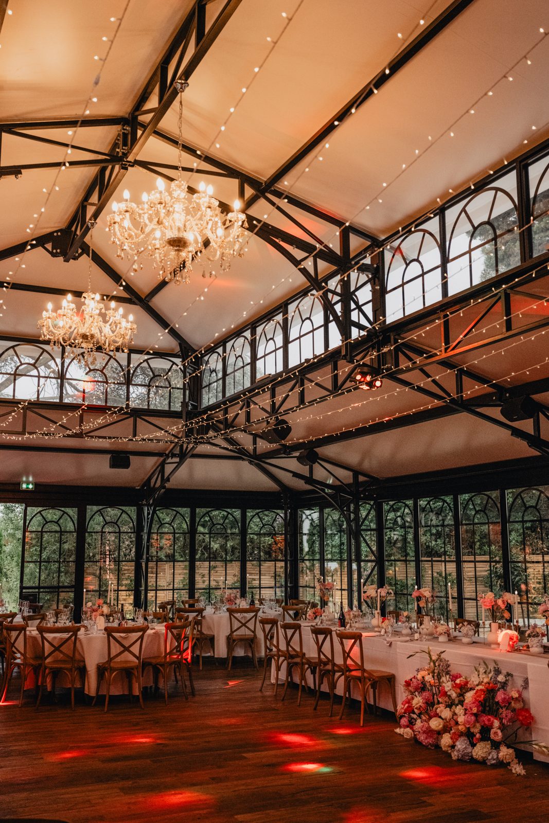 Verrière illuminée de guirlandes lumineuses et de lustres en cristal, tables dressées et bouquet de sol de pivoines