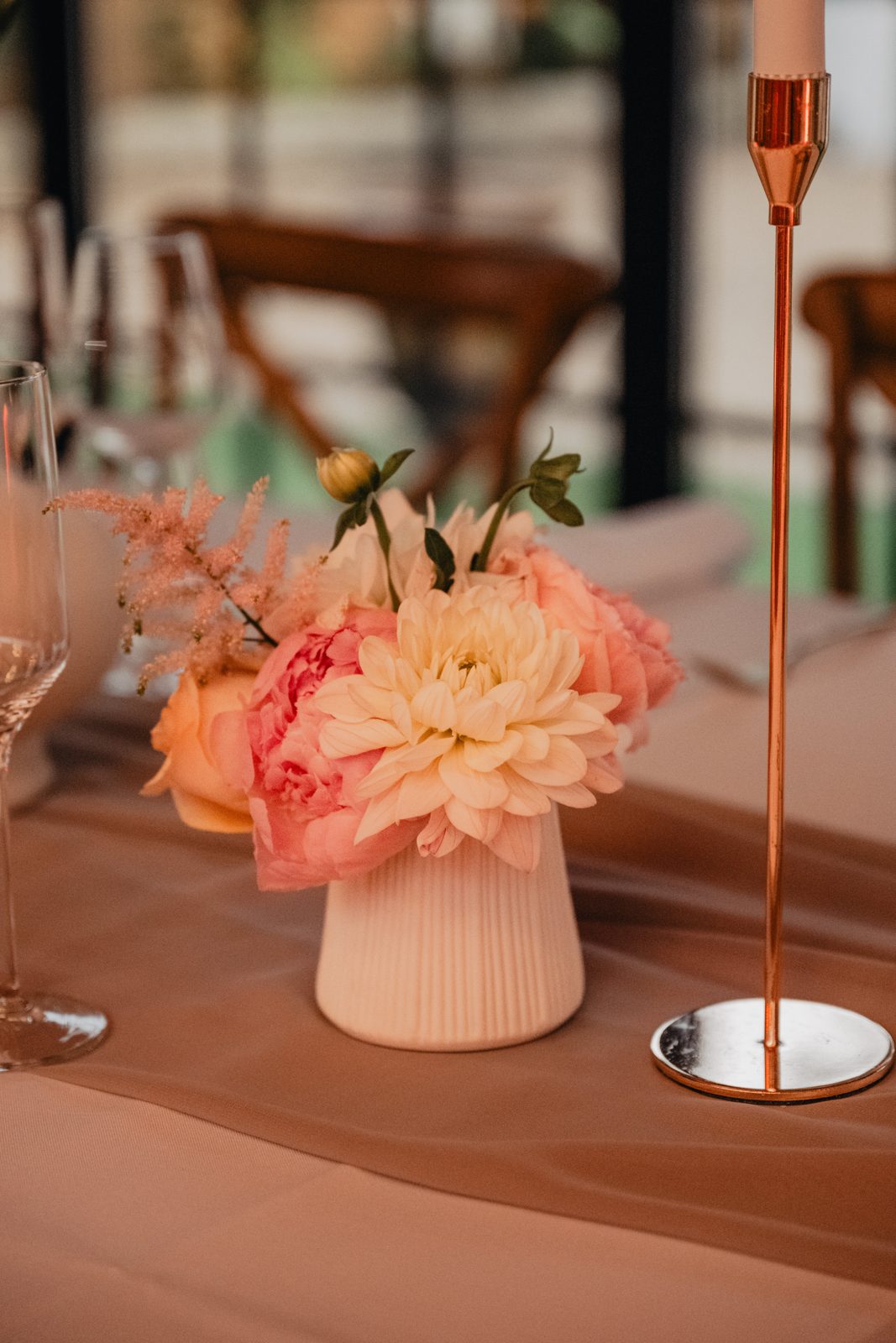 Petit vase cannelé blanc garni de dahlia, pivoine et roses pêche, bougeoir doré sur pied sur un chemin de table rosé