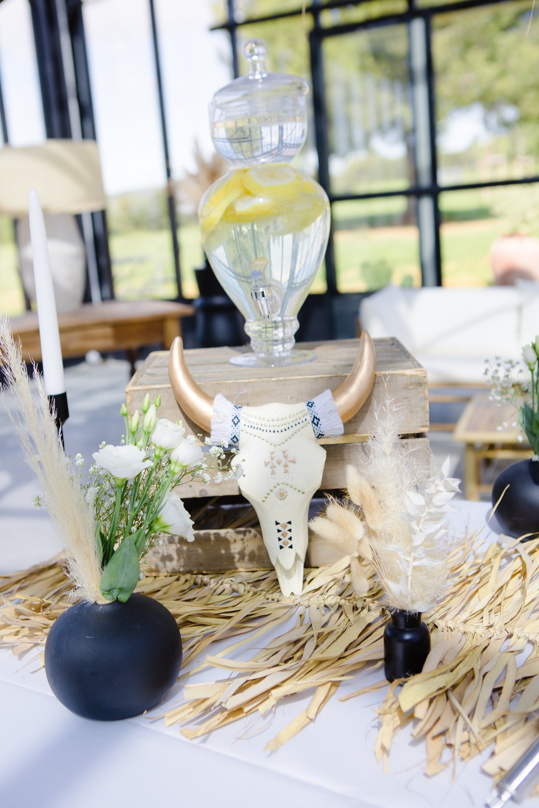Crâne de buffle décoré posé contre une caisse en bois, fontaine à citronnade en verre et petits vases noirs fleuris