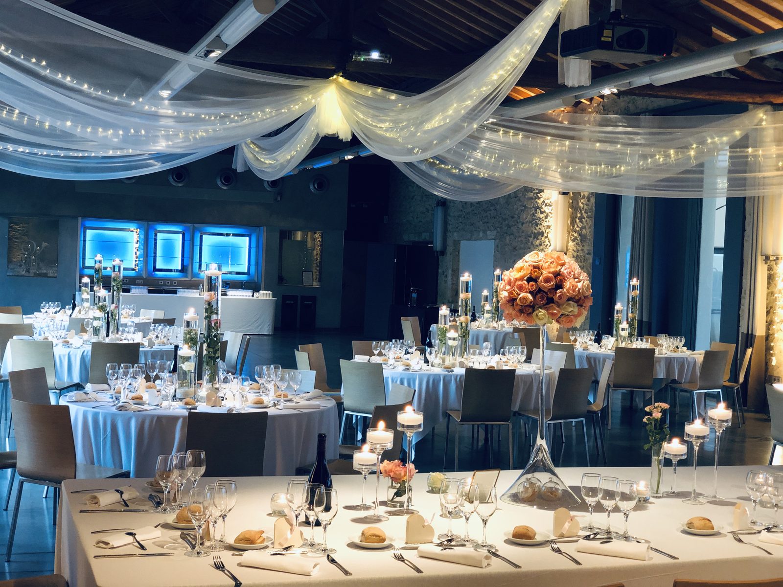 Tables nappées de blanc sous des drapés lumineux, vases cylindres à bougies flottantes et boule de roses sur vase Martini