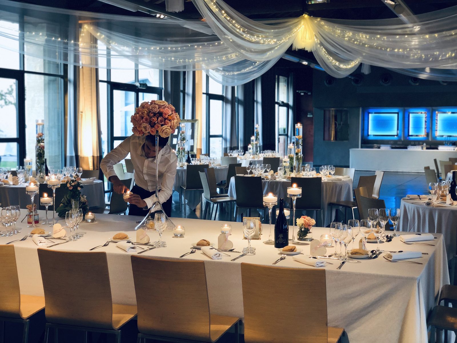 Mise en place d’une table d’honneur avec photophores, vases cylindres fleuris et boule de roses, sous un ciel de voilages