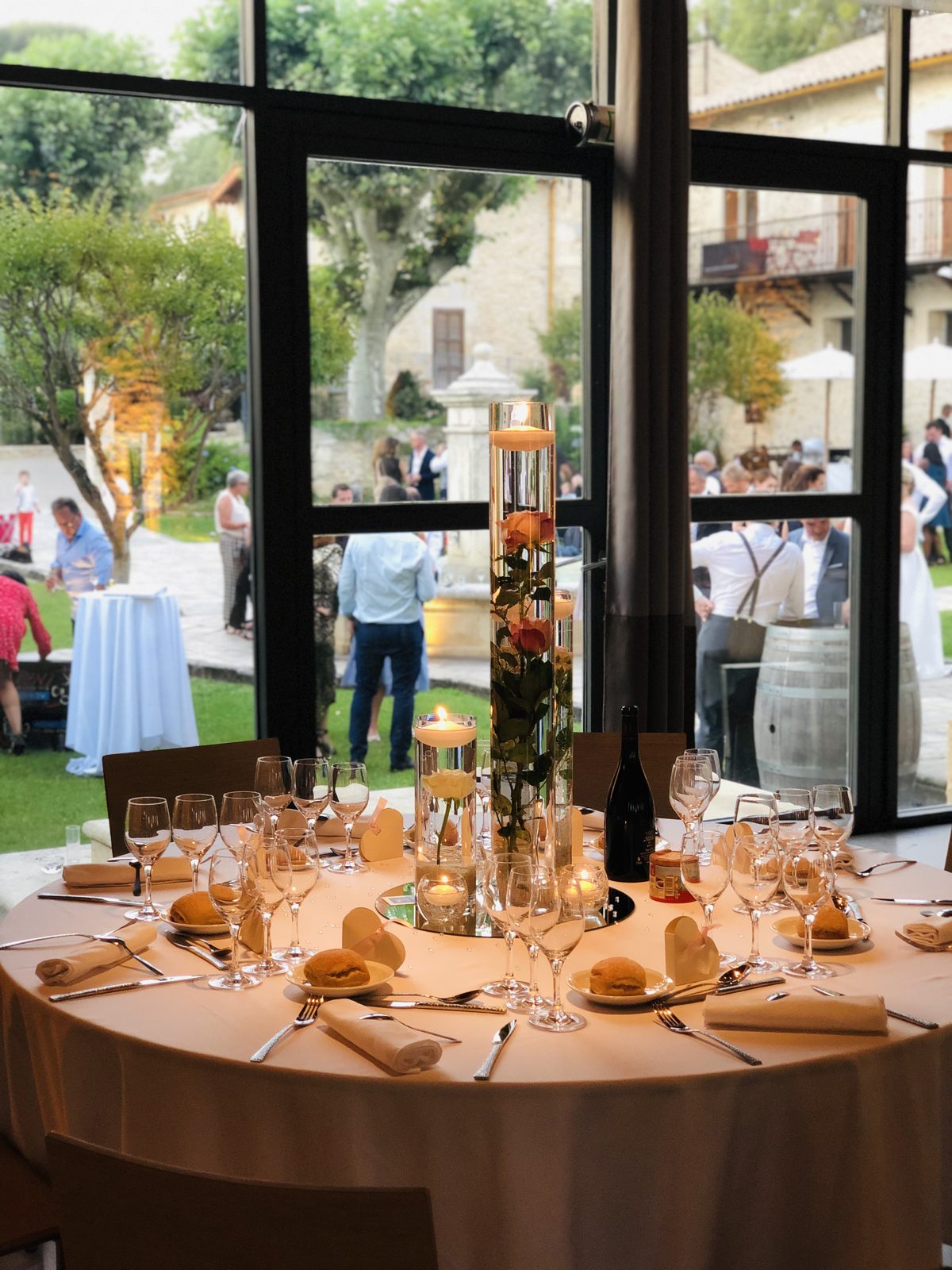Table ronde dressée devant une baie vitrée, vase cylindre avec roses immergées et bougie flottante, cocktail au jardin