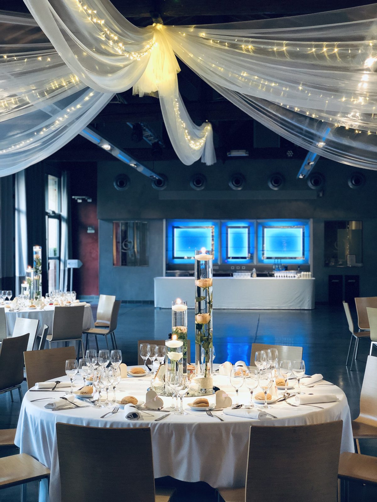 Table ronde nappée de blanc sous des voilages et guirlandes lumineuses, vases cylindres superposés avec bougies flottantes