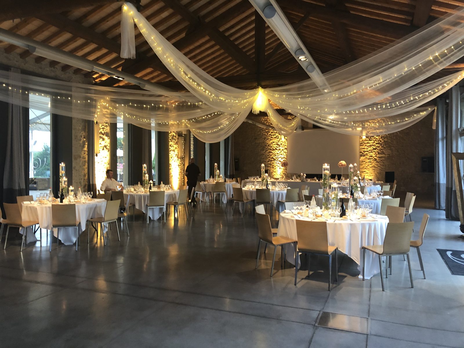 Salle aux murs de pierre et poutres en bois, plafond drapé de voilages blancs et guirlandes, tables rondes nappées de blanc