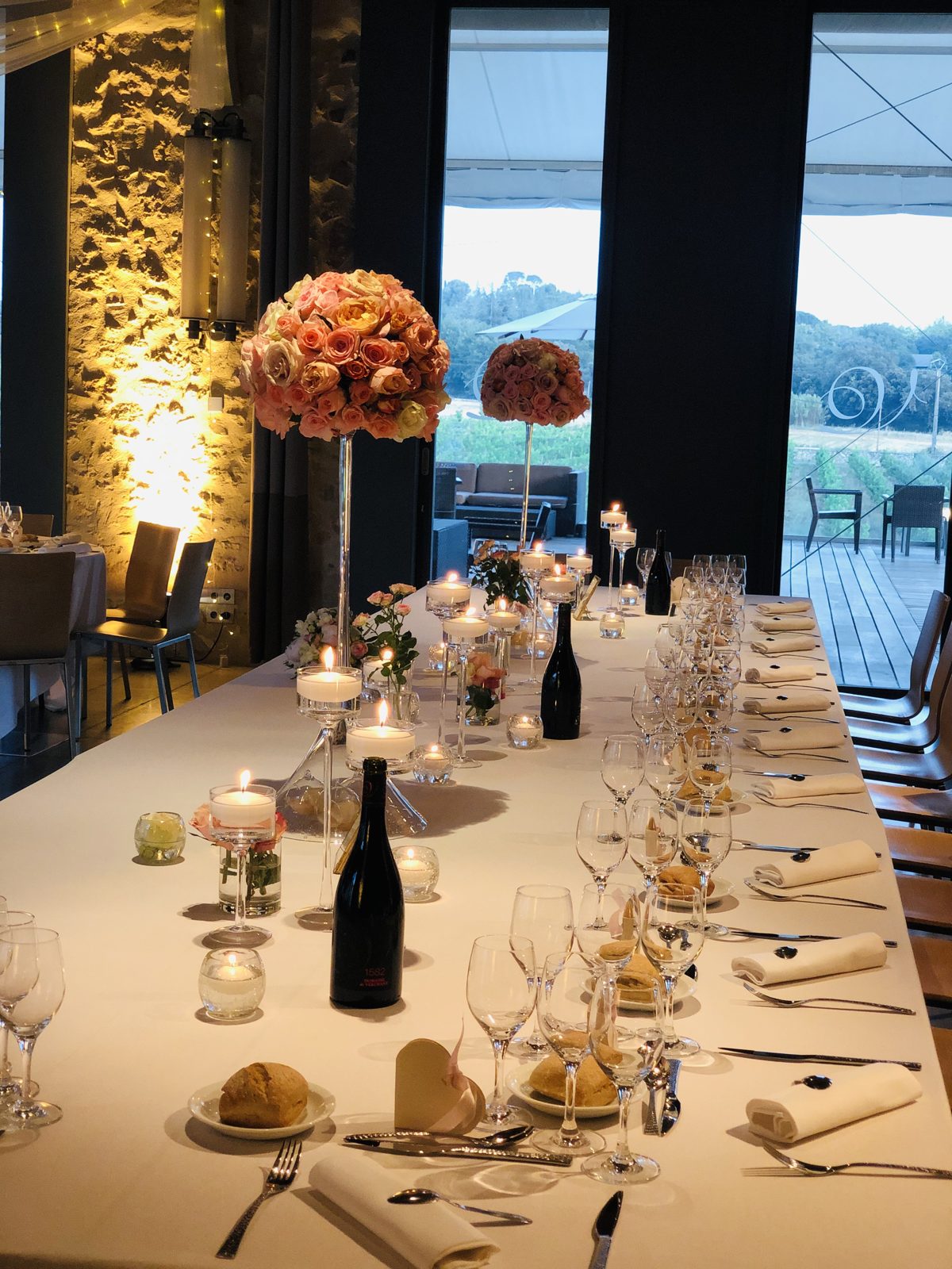 Longue table d’honneur aux nappes écrues, photophores, bougies sur pied et boules de roses sur hauts vases, face aux vignes