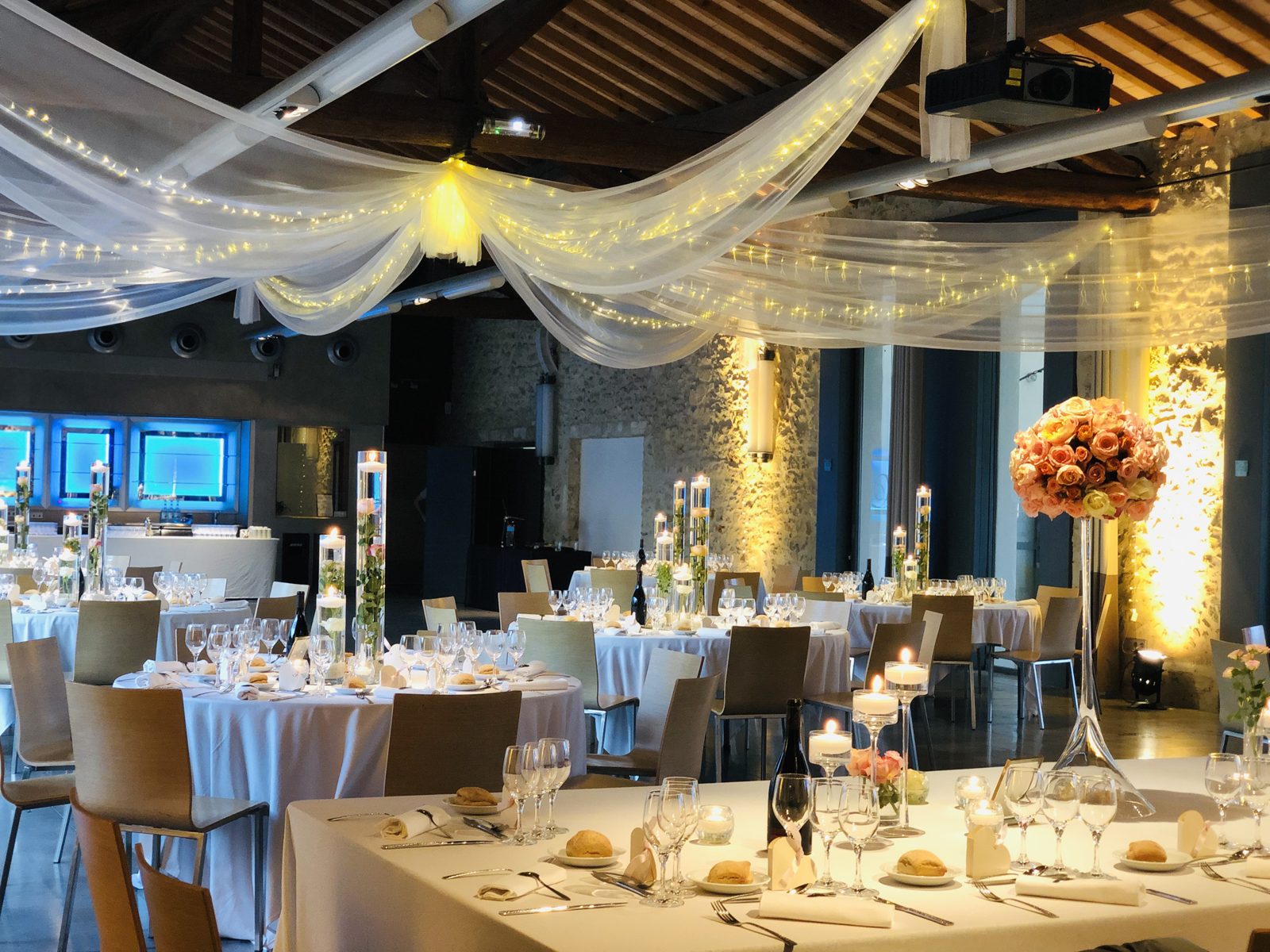 Salle illuminée de voilages et guirlandes, tables rondes aux vases cylindres fleuris et table d’honneur à boule de roses