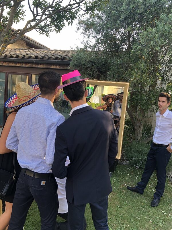 Invités coiffés de chapeaux et lunettes de fête devant une borne photo miroir au cadre doré lumineux, dans le jardin d’un domaine