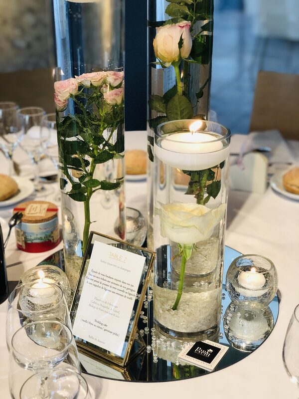 Centre de table sur miroir rond : vases cylindres aux roses immergées et bougie flottante, photophores en verre craquelé, menu dans un cadre doré
