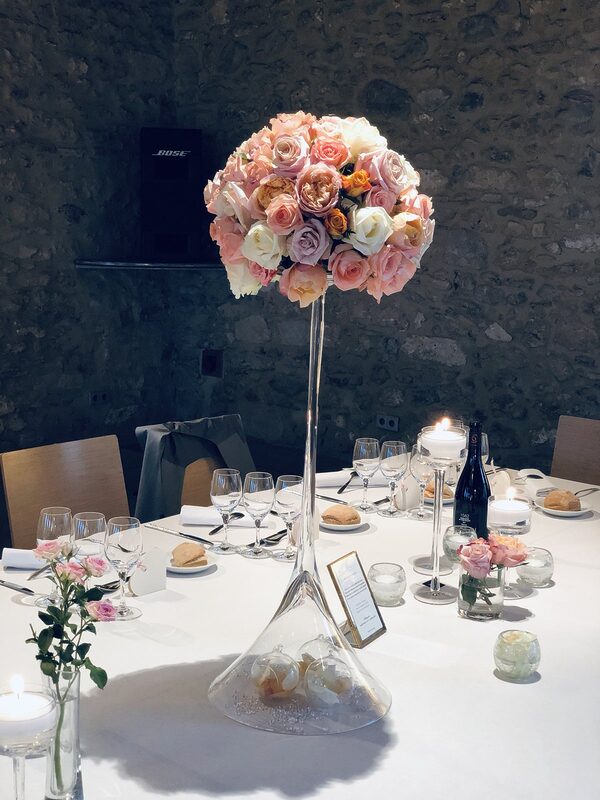 Boule de roses pêche, roses et blanches sur un haut vase Martini, bougies flottantes sur pied, devant un mur de pierre