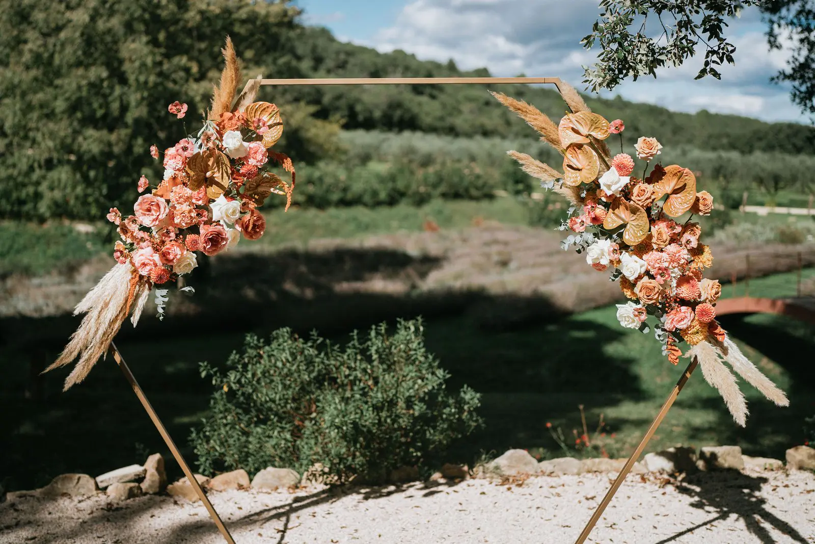 Arche hexagonale dorée fleurie de roses, anthuriums et pampas aux tons terracotta, devant un jardin et une oliveraie