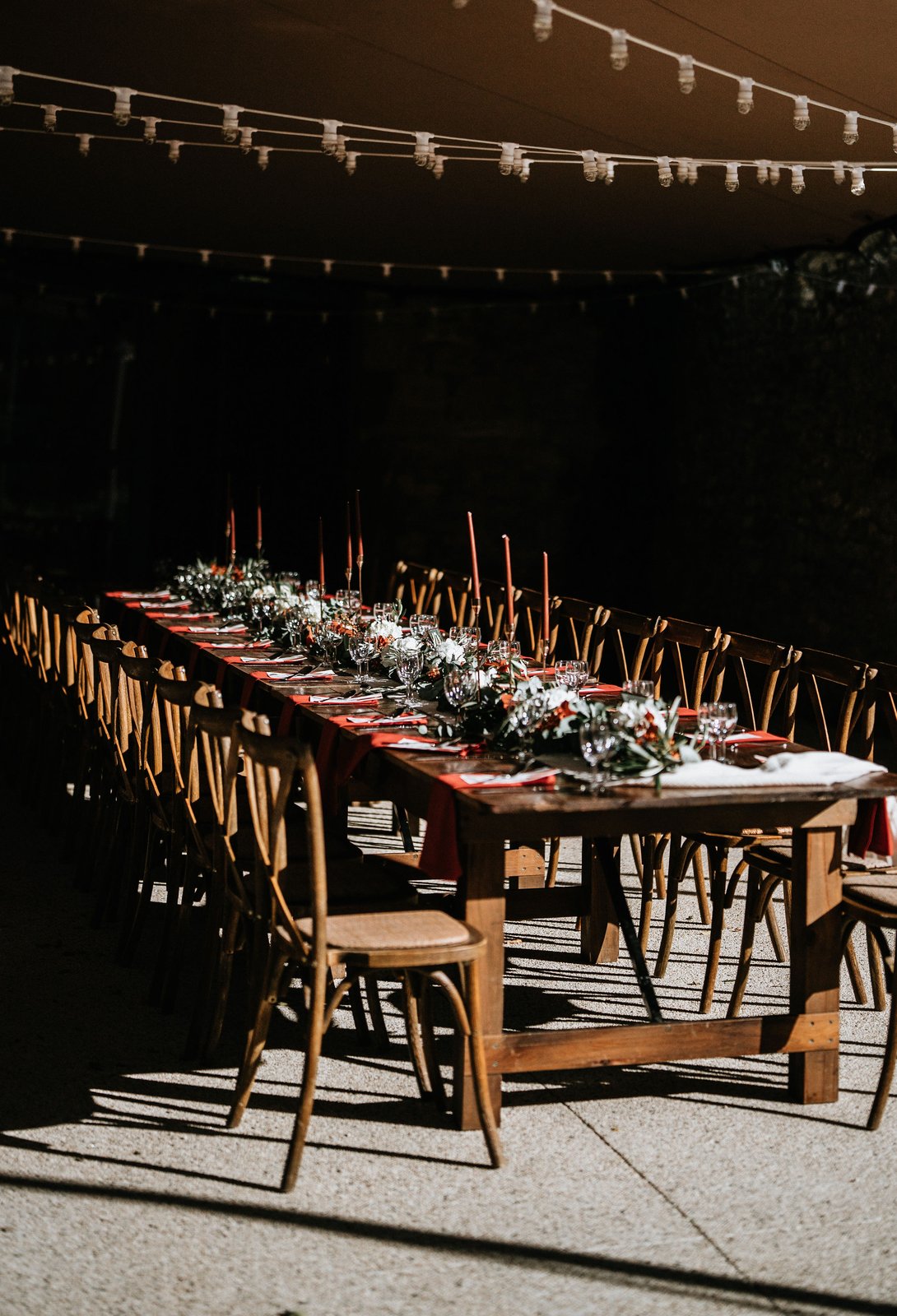 Longue table en bois sous une tente tendue éclairée de guirlandes, chaises bistrot en bois et chandelles bordeaux