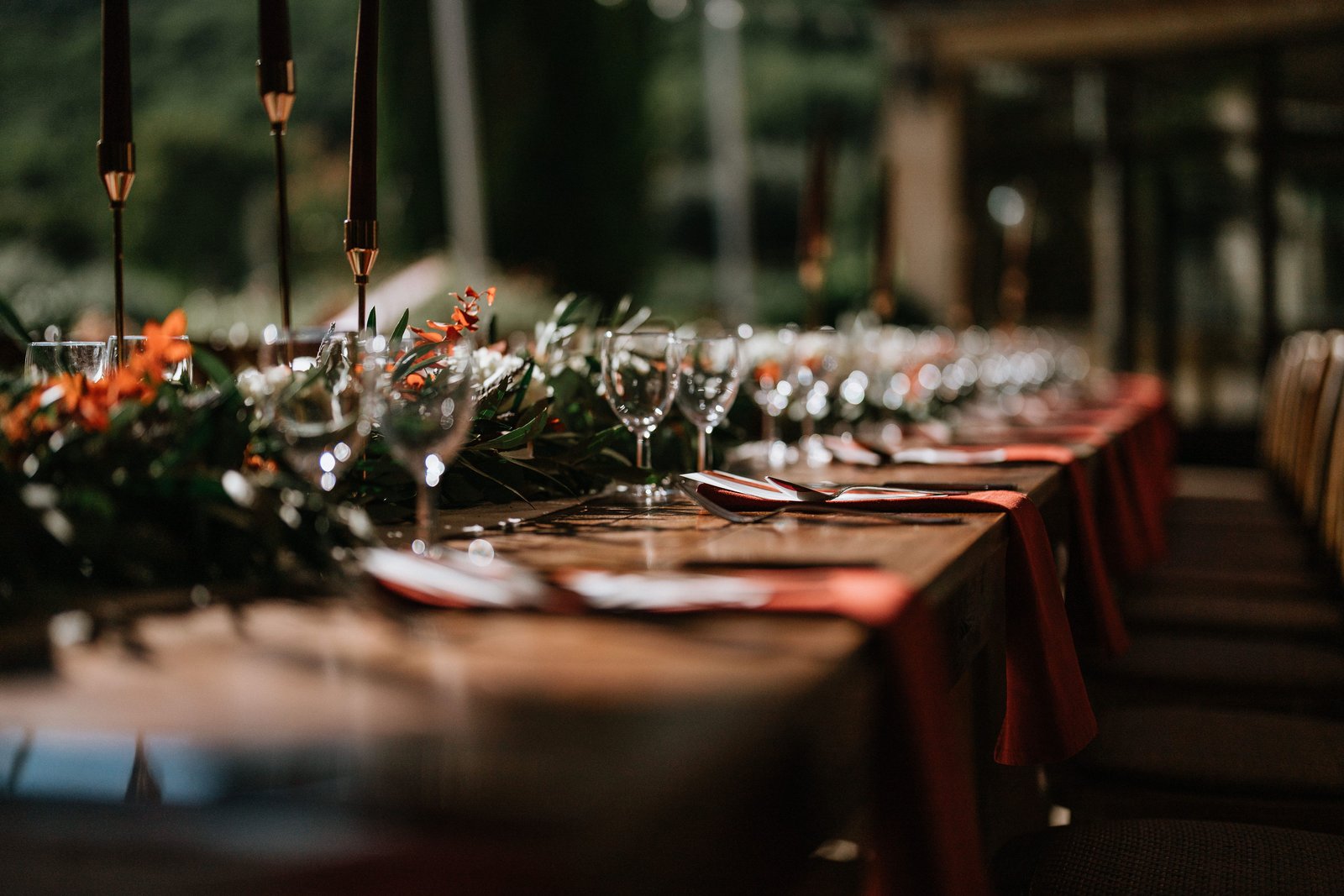 Table en bois dressée de verres, serviettes rouges et guirlande de feuillage et fleurs orangées, chandelles bordeaux