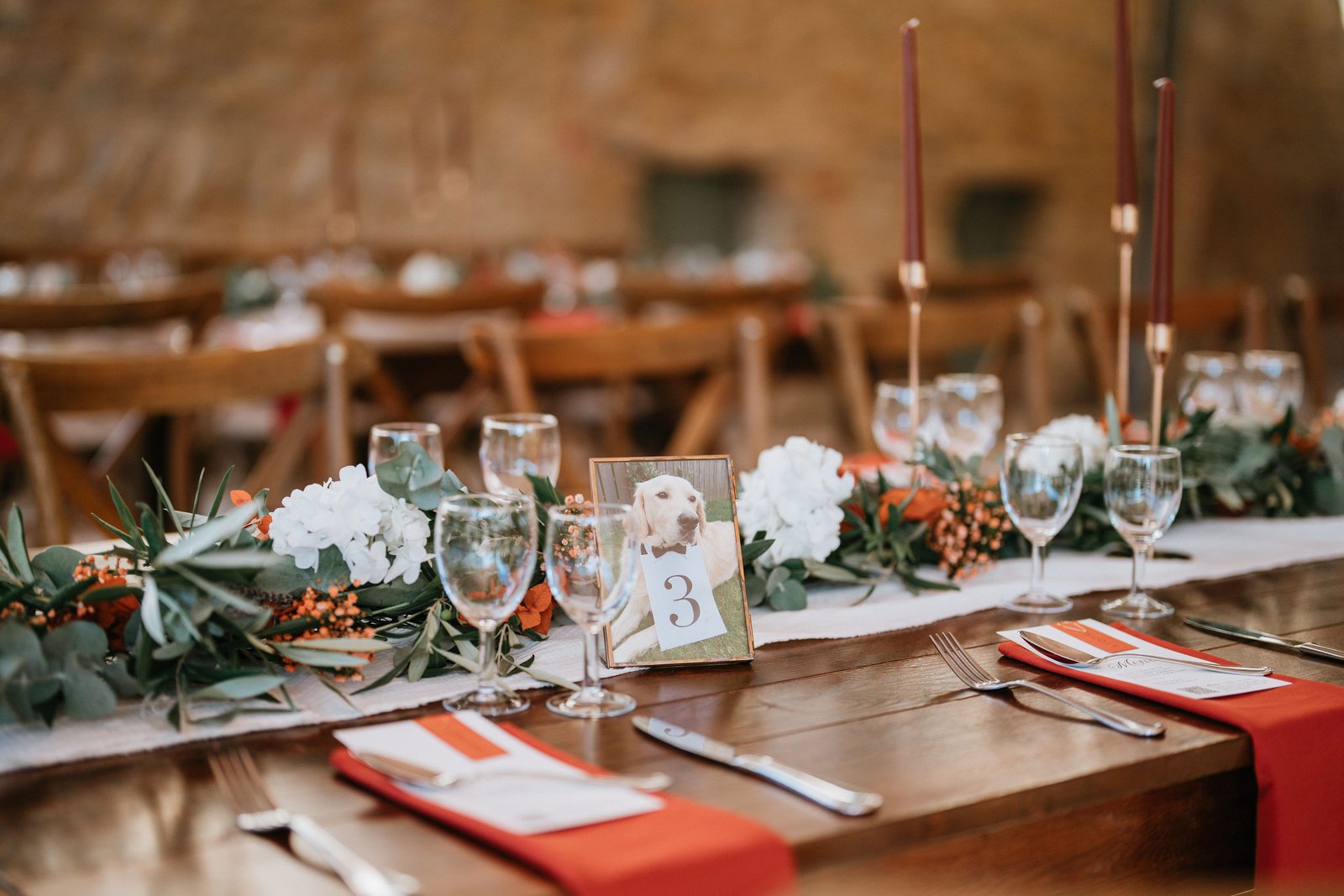 Guirlande d’eucalyptus et d’hortensias blancs sur chemin de gaze, numéro de table encadré, serviettes rouges sur table en bois