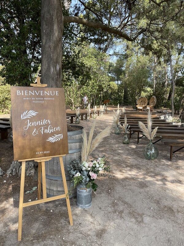 Panneau de bienvenue en bois sur chevalet au pied d’un tonneau, bidon en zinc fleuri, allée de bancs en bois sous les pins