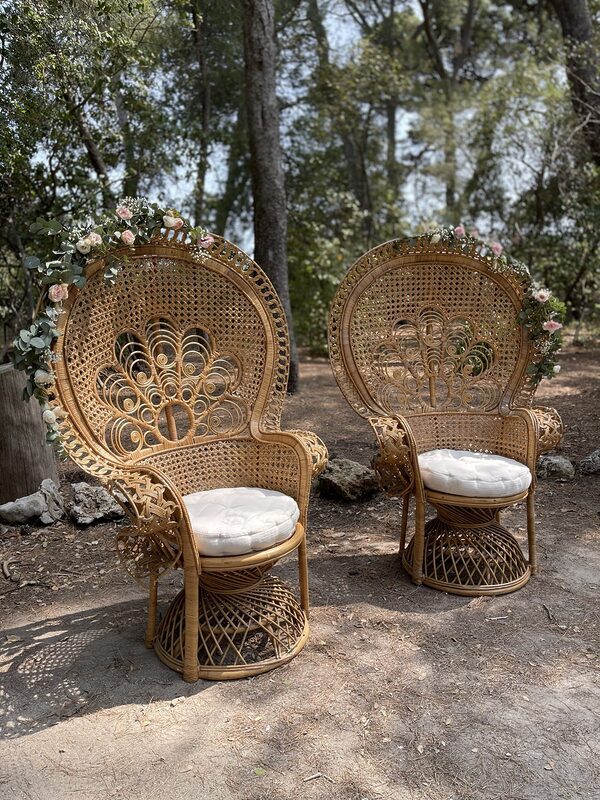 Deux fauteuils paon en rotin aux coussins blancs, guirlandes d’eucalyptus et de roses pâles, dans un sous-bois