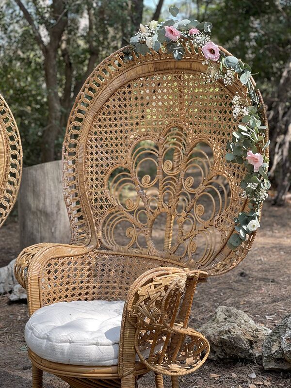 Gros plan sur un fauteuil paon en rotin, dossier ajouré orné d’une guirlande de fleurs roses et d’eucalyptus