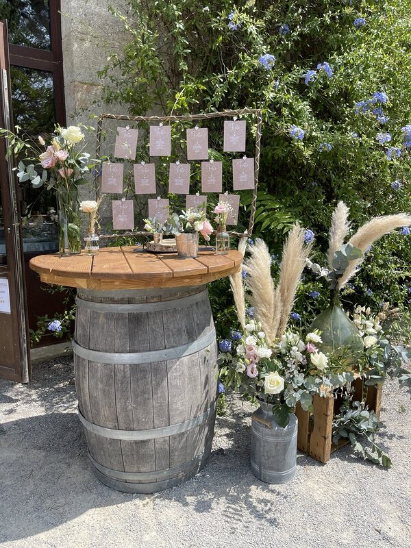 Plan de table sur un tonneau mange-debout à plateau en bois, bidon en zinc et dame-jeanne garnis de pampas, roses et eucalyptus, caisse en bois