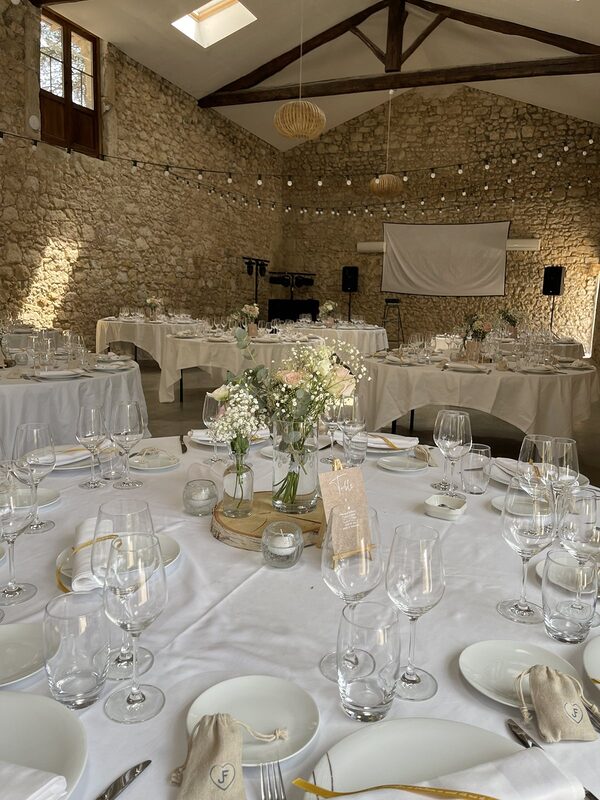 Salle aux murs de pierre : guirlande guinguette, suspensions en rotin, tables rondes nappées de blanc, centres de table sur rondin de bois