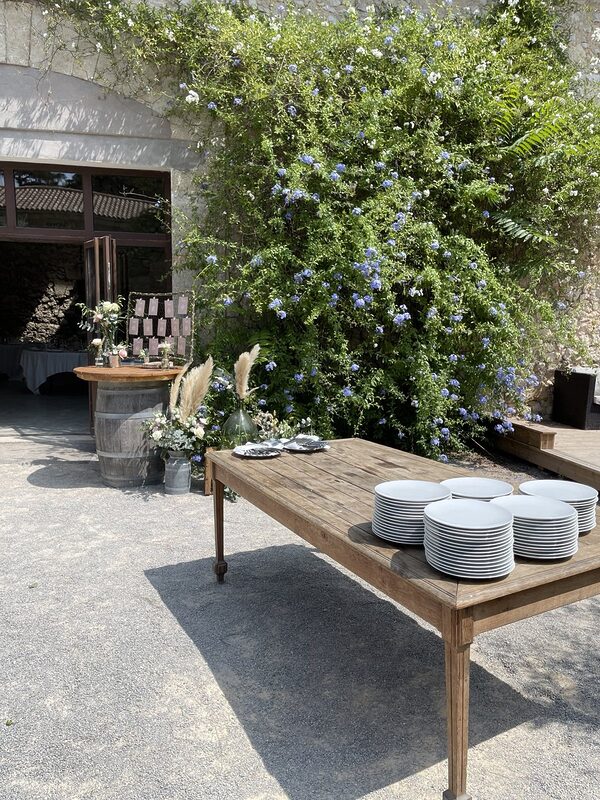 Cour du domaine sous un plumbago en fleurs, tonneau du plan de table et compositions de pampas à l’entrée de la salle