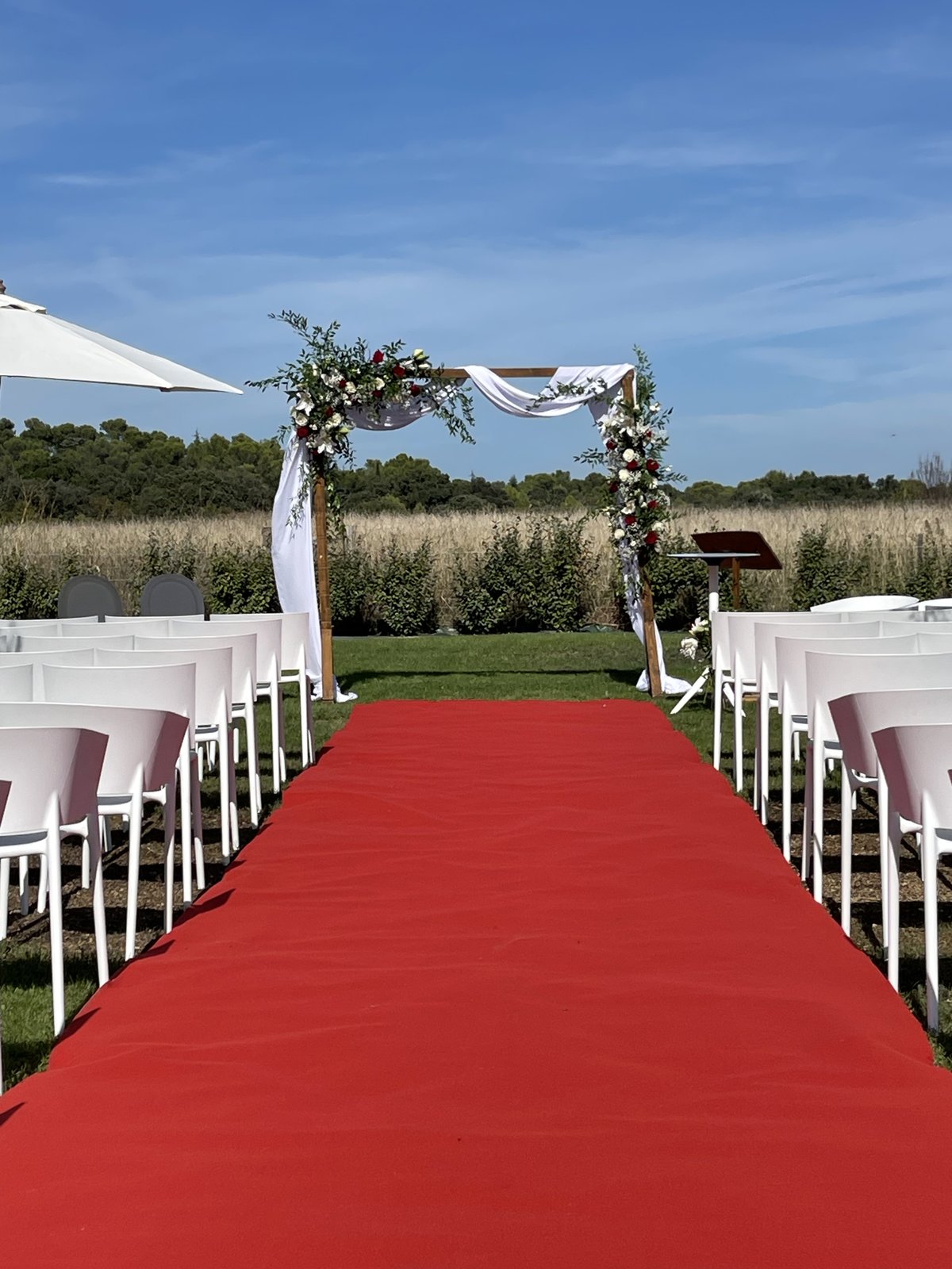 Allée de cérémonie en moquette rouge entre des chaises blanches, vers une arche en bois drapée de blanc et fleurie