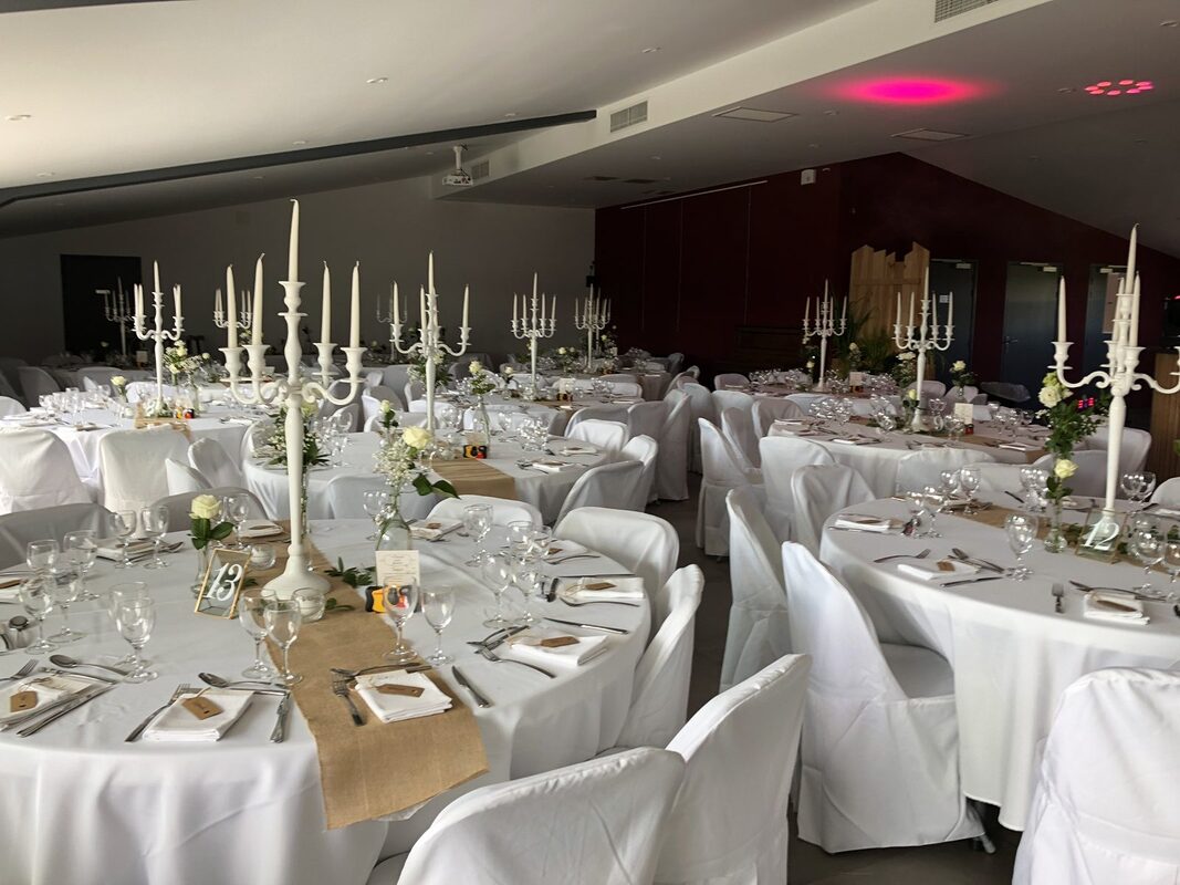Salle de réception aux tables rondes nappées de blanc, grands chandeliers laqués blancs à cinq branches, chemins de jute et chaises housées de blanc