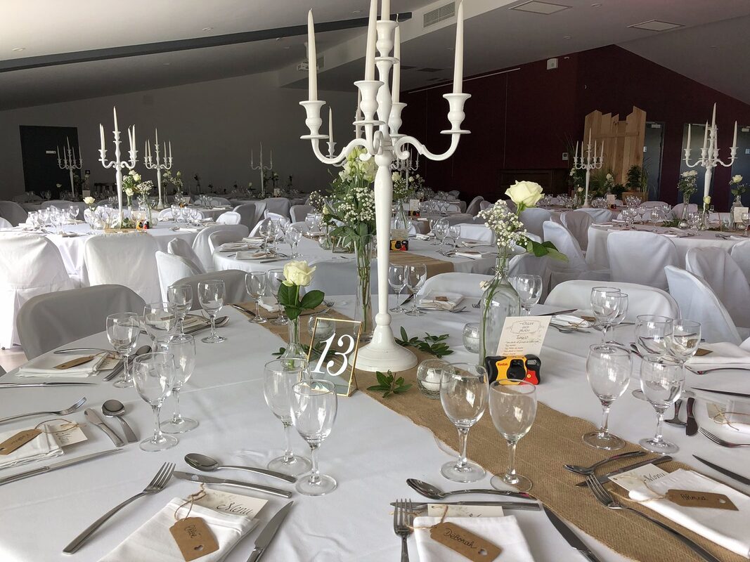 Centre de table : chandelier laqué blanc, roses blanches en soliflore, numéro de table dans un cadre doré, chemin de jute et photophores en verre craqué