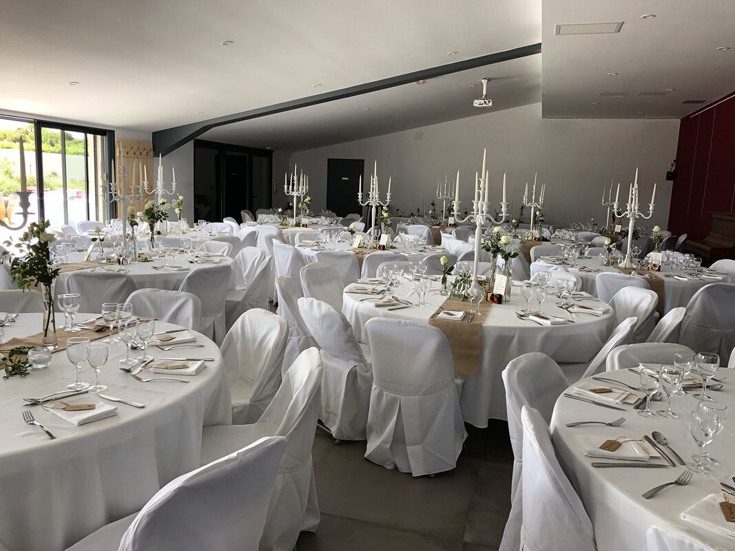 Vue d’ensemble de la salle : chandeliers blancs sur chaque table, chaises housées de blanc, baie vitrée ouverte sur le jardin
