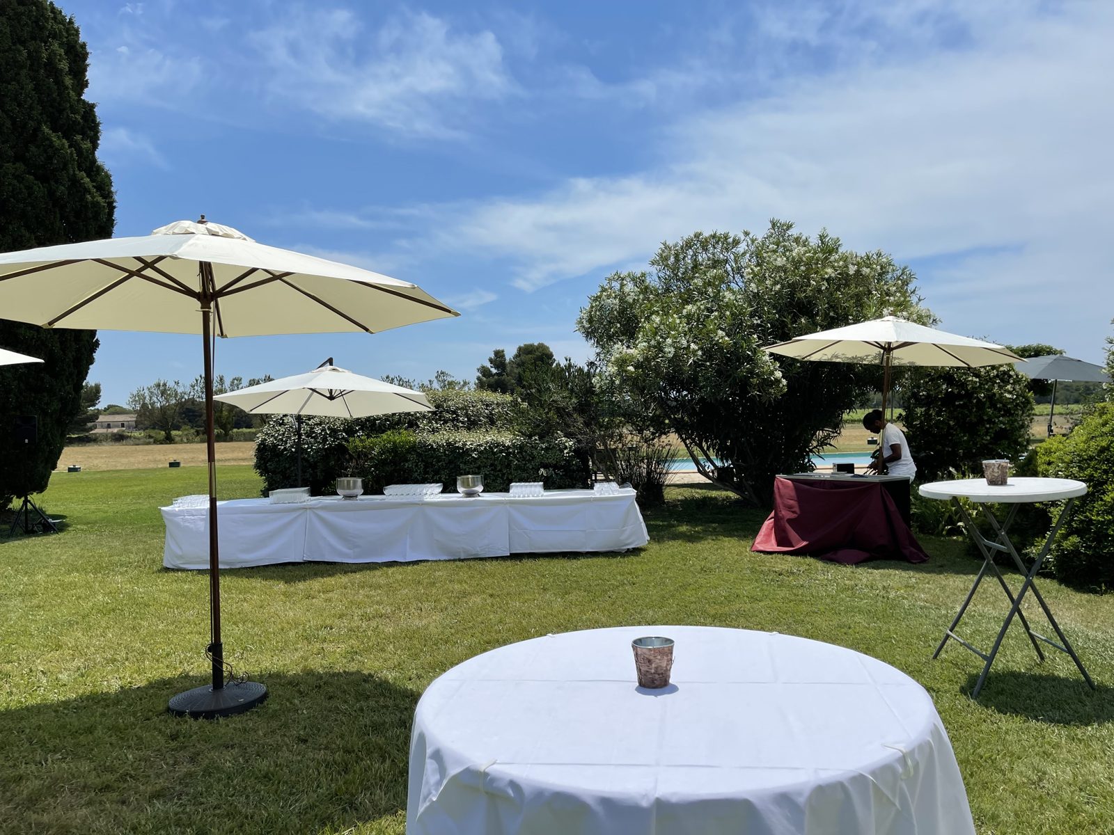 Cocktail sur la pelouse avec parasols écrus, buffet nappé de blanc et mange-debout recouverts de nappes blanches