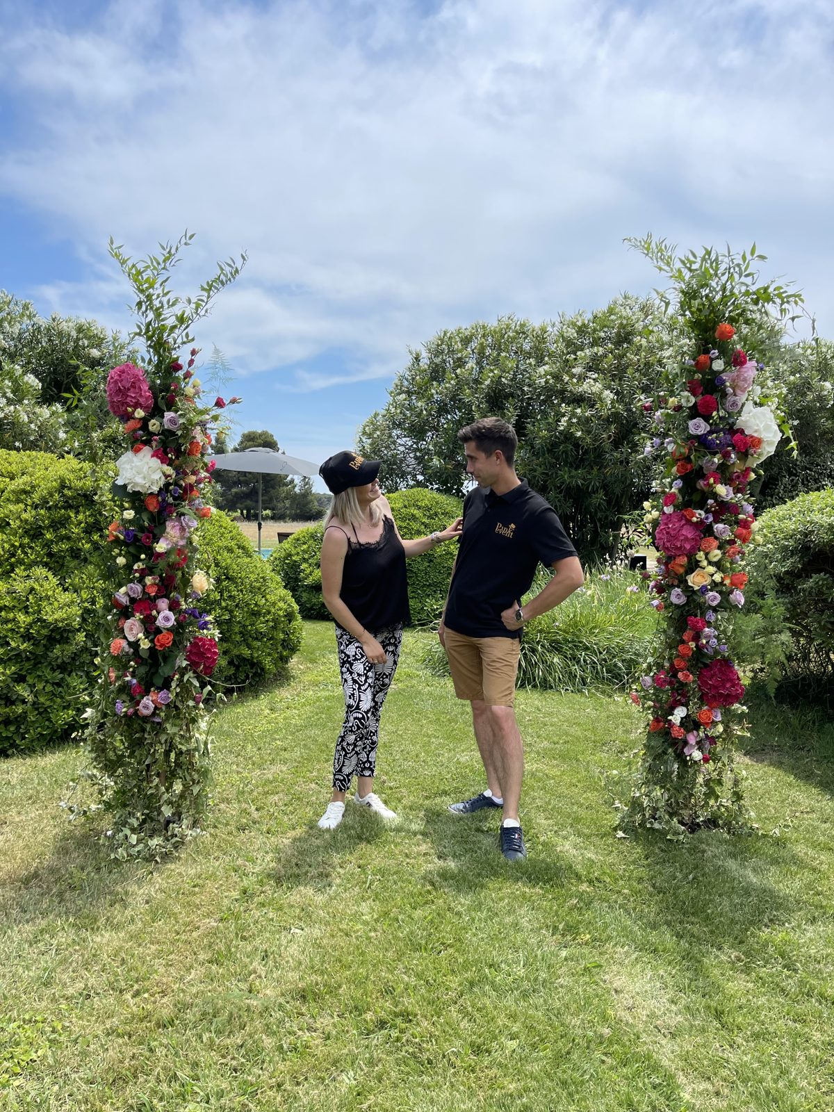 Deux colonnes florales aux fleurs rouges, roses et violettes dressées sur la pelouse d’un jardin, deux personnes au centre