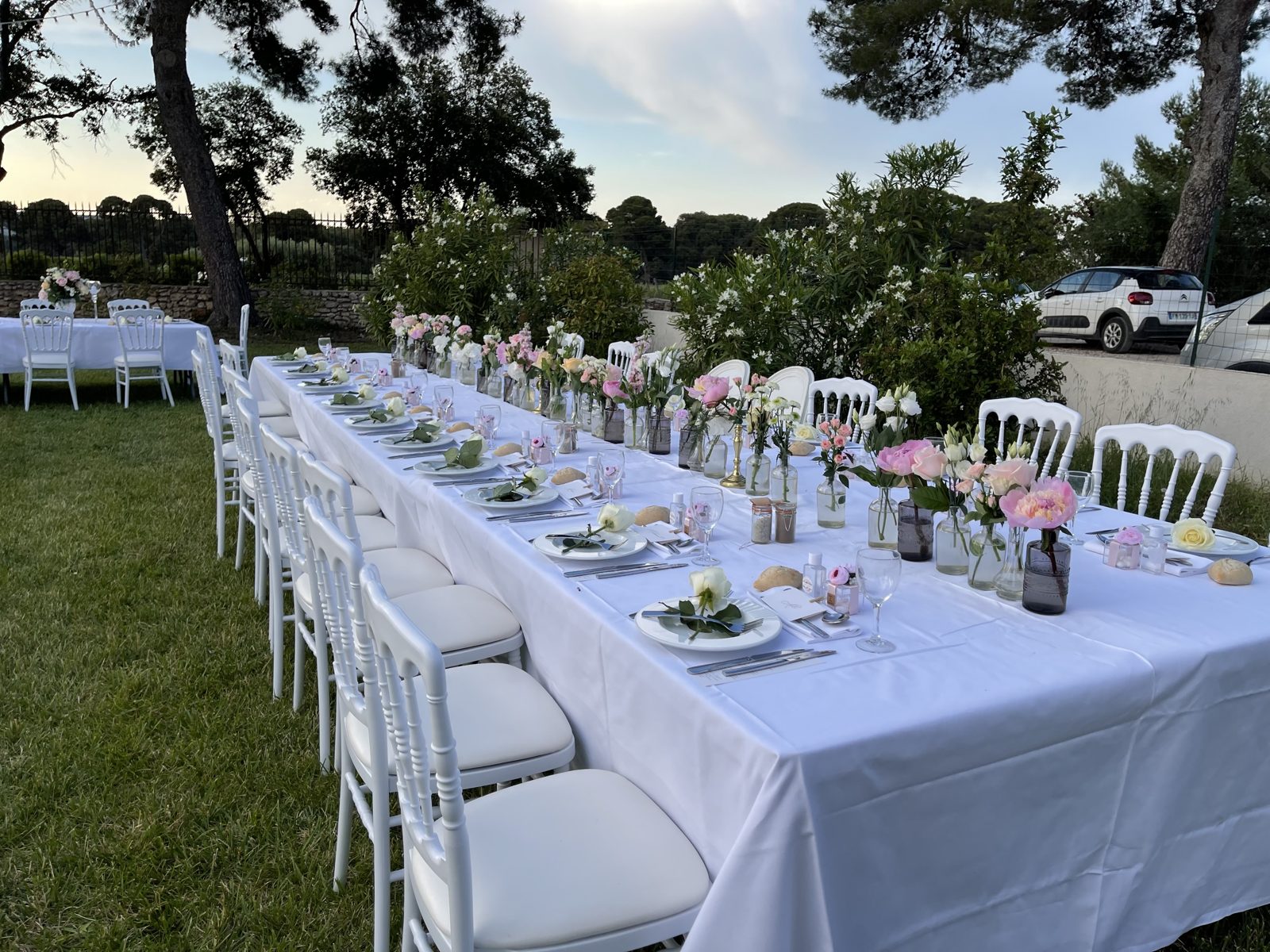 Longue table nappée de blanc au jardin, chaises Napoléon III blanches et petits vases de fleurs roses et blanches