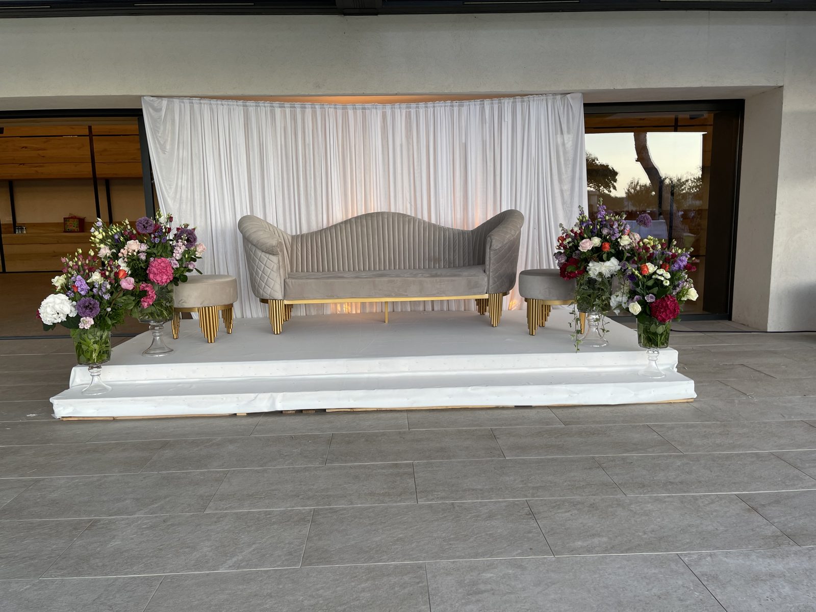 Coin des mariés sur estrade blanche, canapé gris aux pieds dorés devant un rideau blanc plissé, vases de fleurs colorées