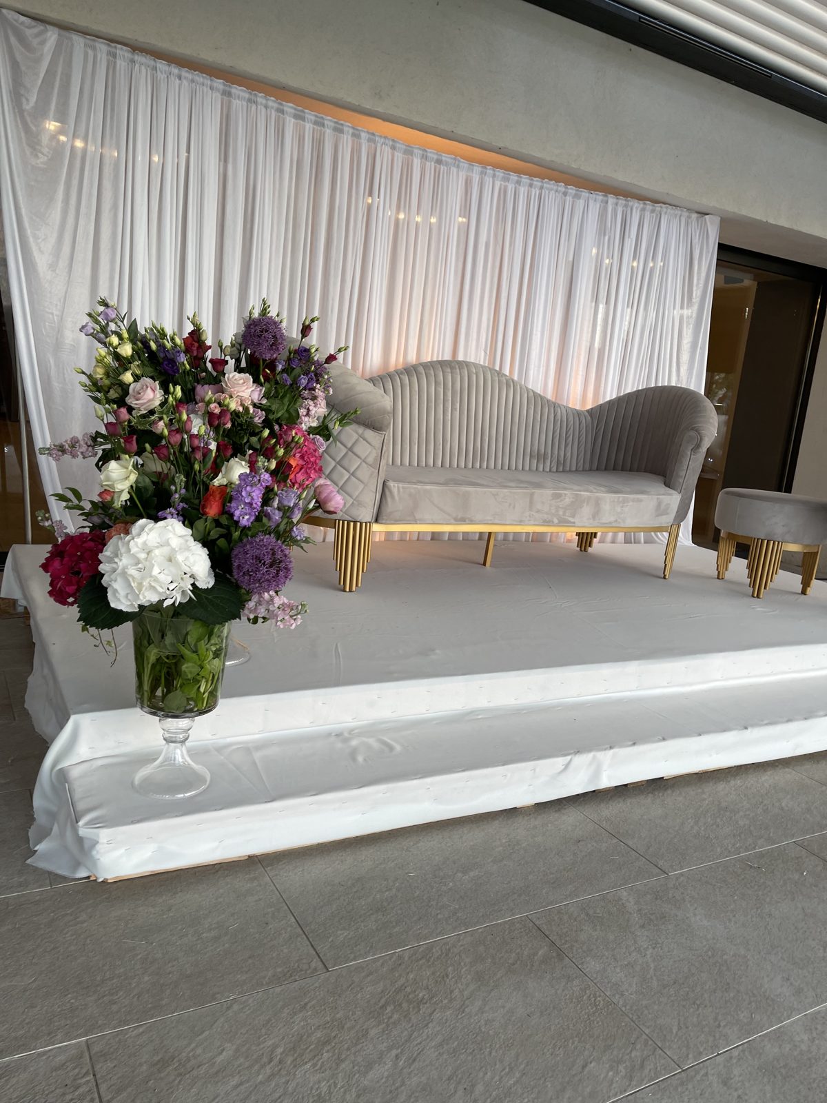 Canapé gris velours et pouf aux pieds dorés sur estrade blanche devant un rideau blanc, grand bouquet dans un vase sur pied