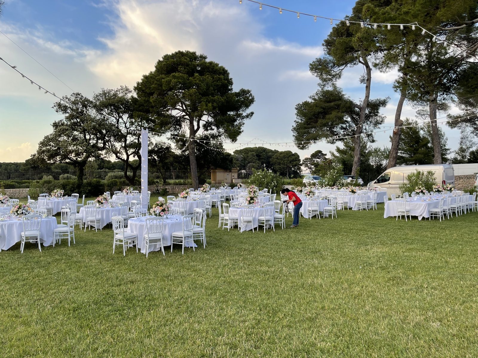 Réception en plein air, tables rondes nappées de blanc, chaises Napoléon III blanches et guirlande guinguette entre les pins