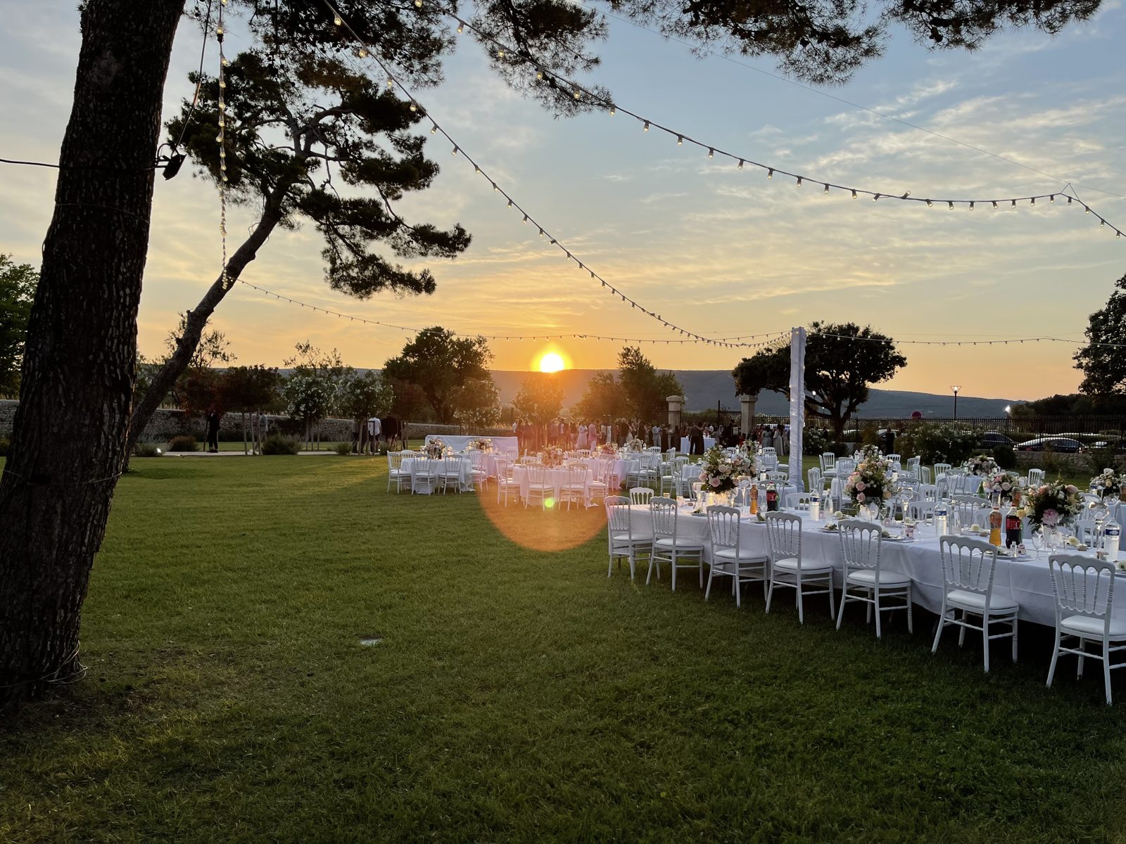 Dîner au jardin au soleil couchant, longues tables blanches, chaises Napoléon III et guirlande guinguette entre les pins
