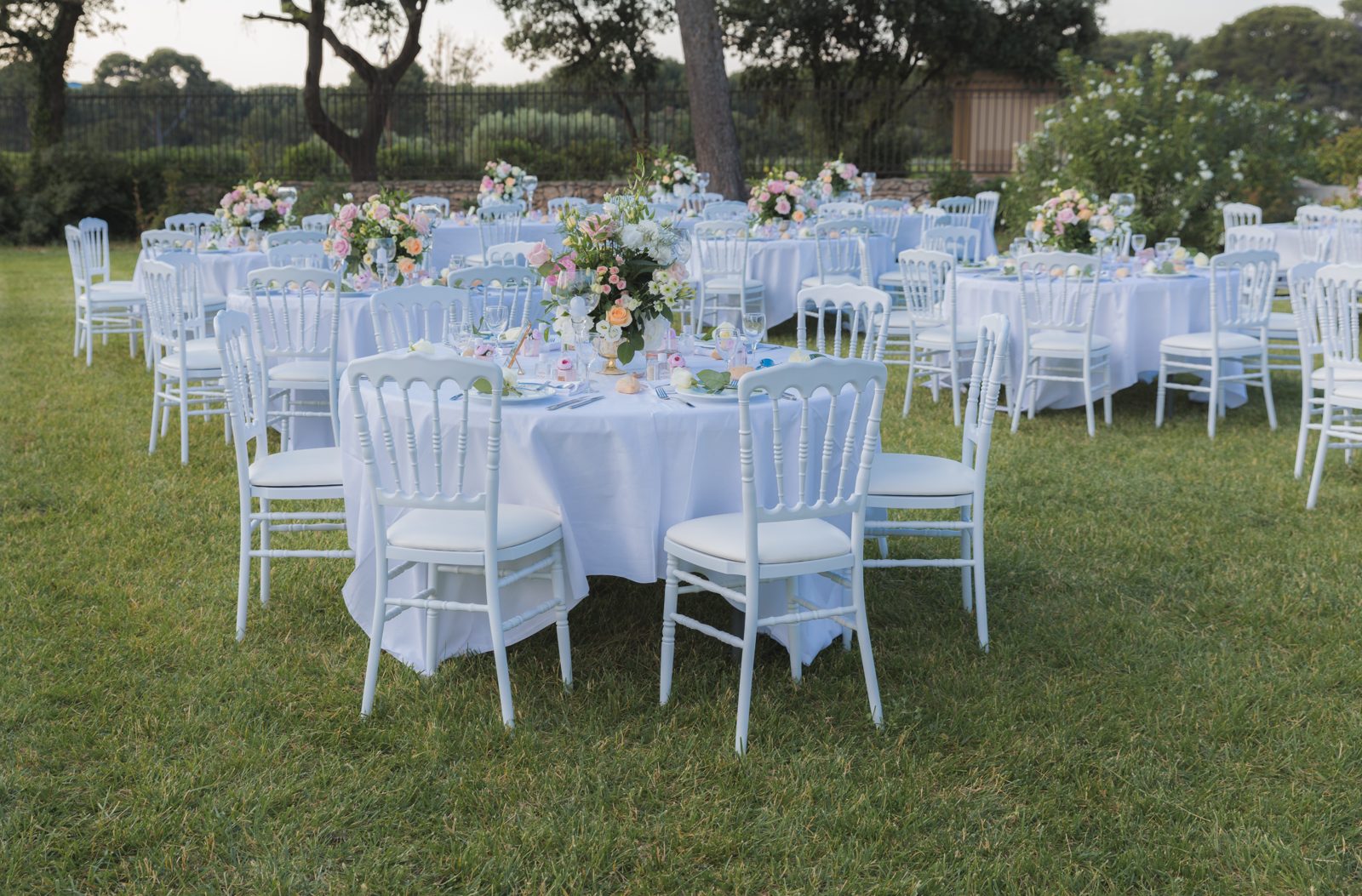 Tables rondes nappées de blanc sur la pelouse, chaises Napoléon III blanches et centres de table aux fleurs roses et pêche