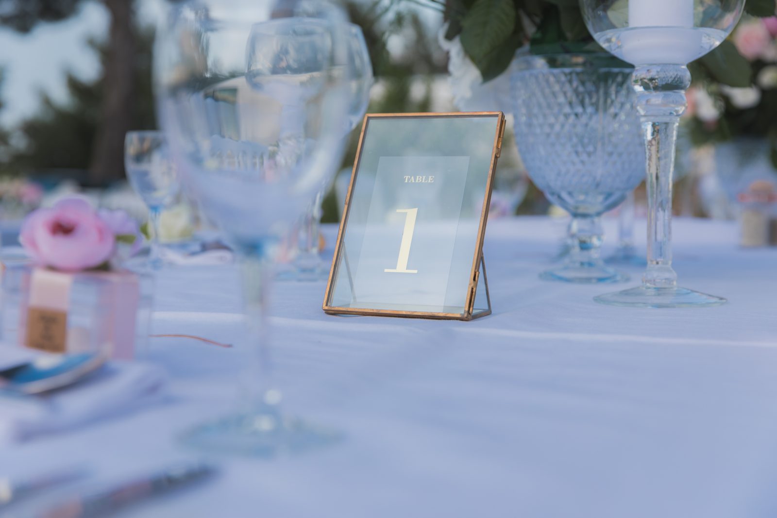 Numéro de table dans un cadre transparent au contour doré, posé sur une nappe blanche entre verres et vase en verre ciselé