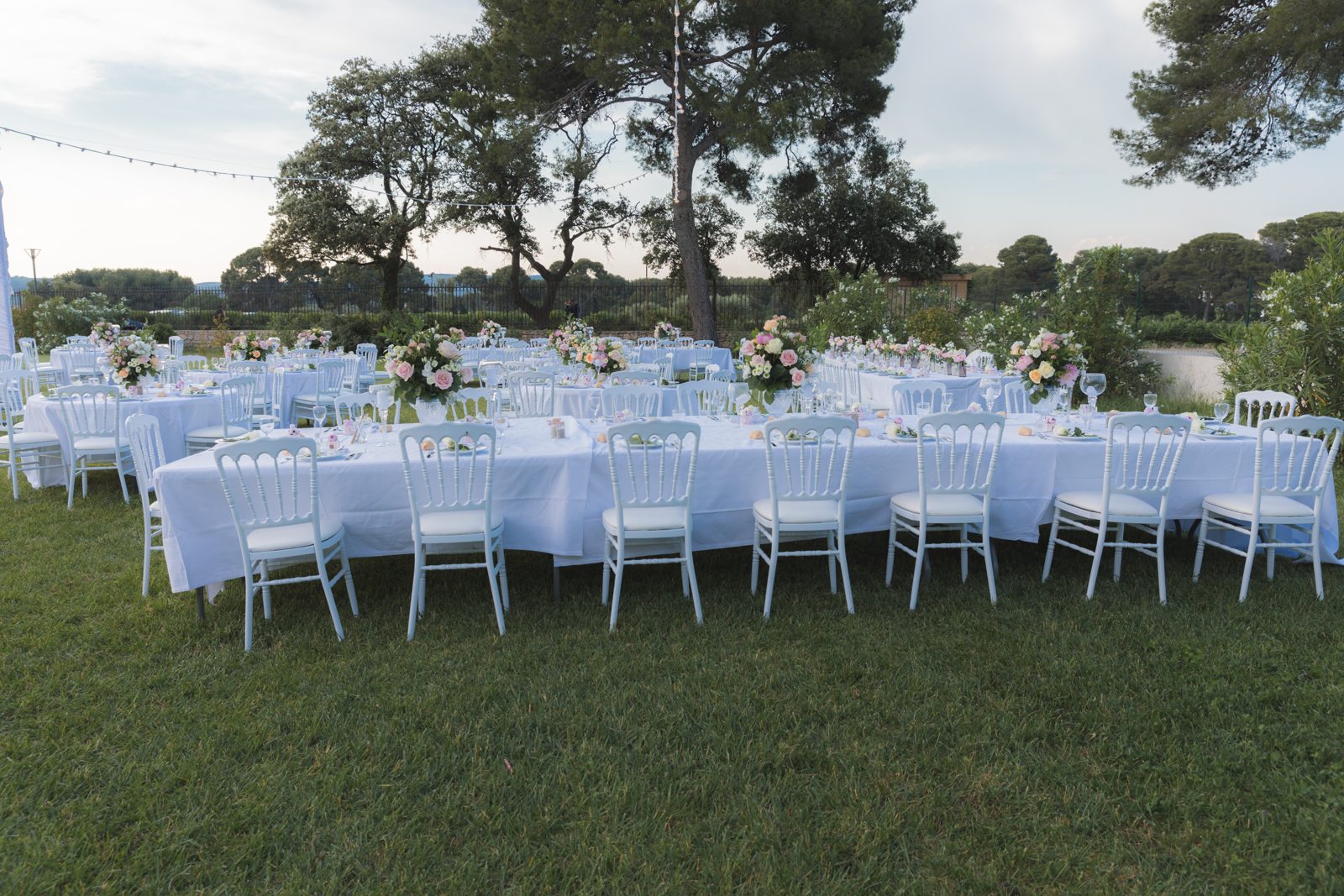 Longue table nappée de blanc et chaises Napoléon III blanches au jardin, centres de table fleuris de roses pâles