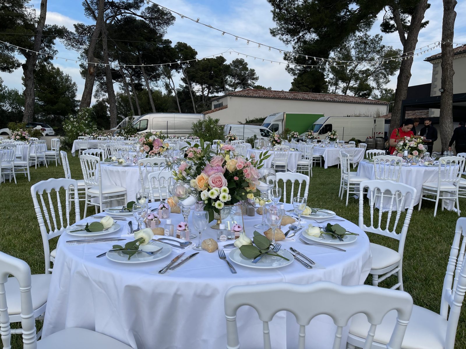 Table ronde dressée au jardin, bouquet de roses pêche et roses dans un vase doré, chaises Napoléon III blanches autour