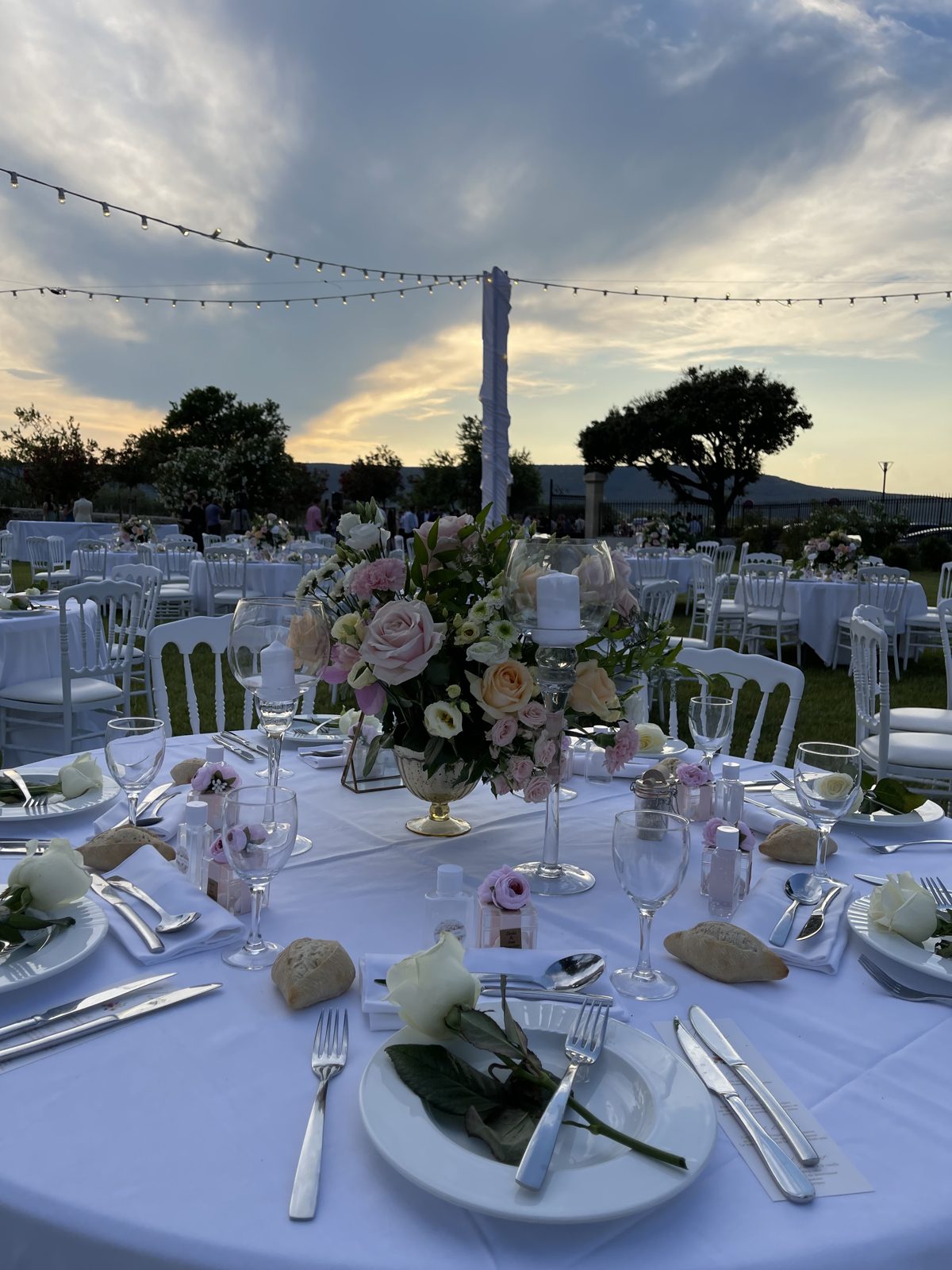 Centre de table fleuri dans un vase compotier doré, bougie sur pied en verre et roses blanches sur les assiettes