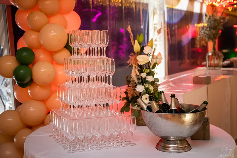 Pyramide de coupes à champagne et seau à bouteilles sur table nappée, arche de ballons pêche et verts sous chapiteau