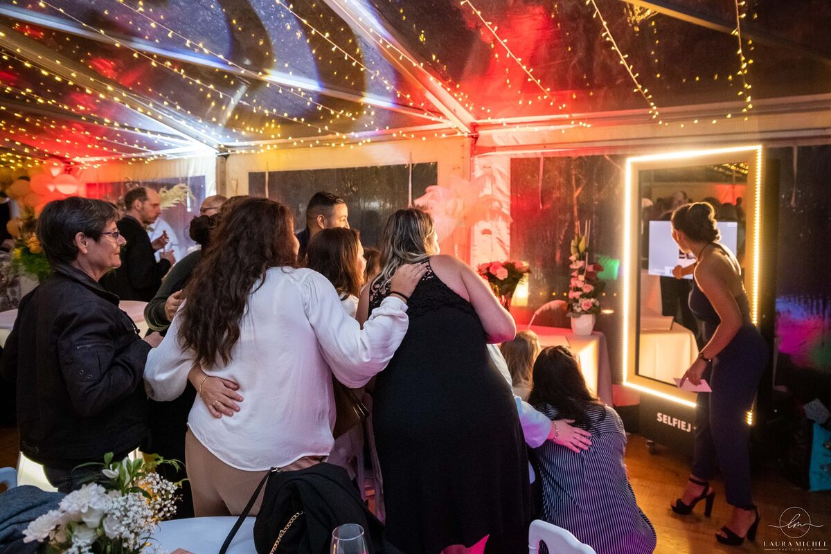 Invités devant une borne photo miroir encadrée de lumières, sous un chapiteau transparent tendu de guirlandes lumineuses