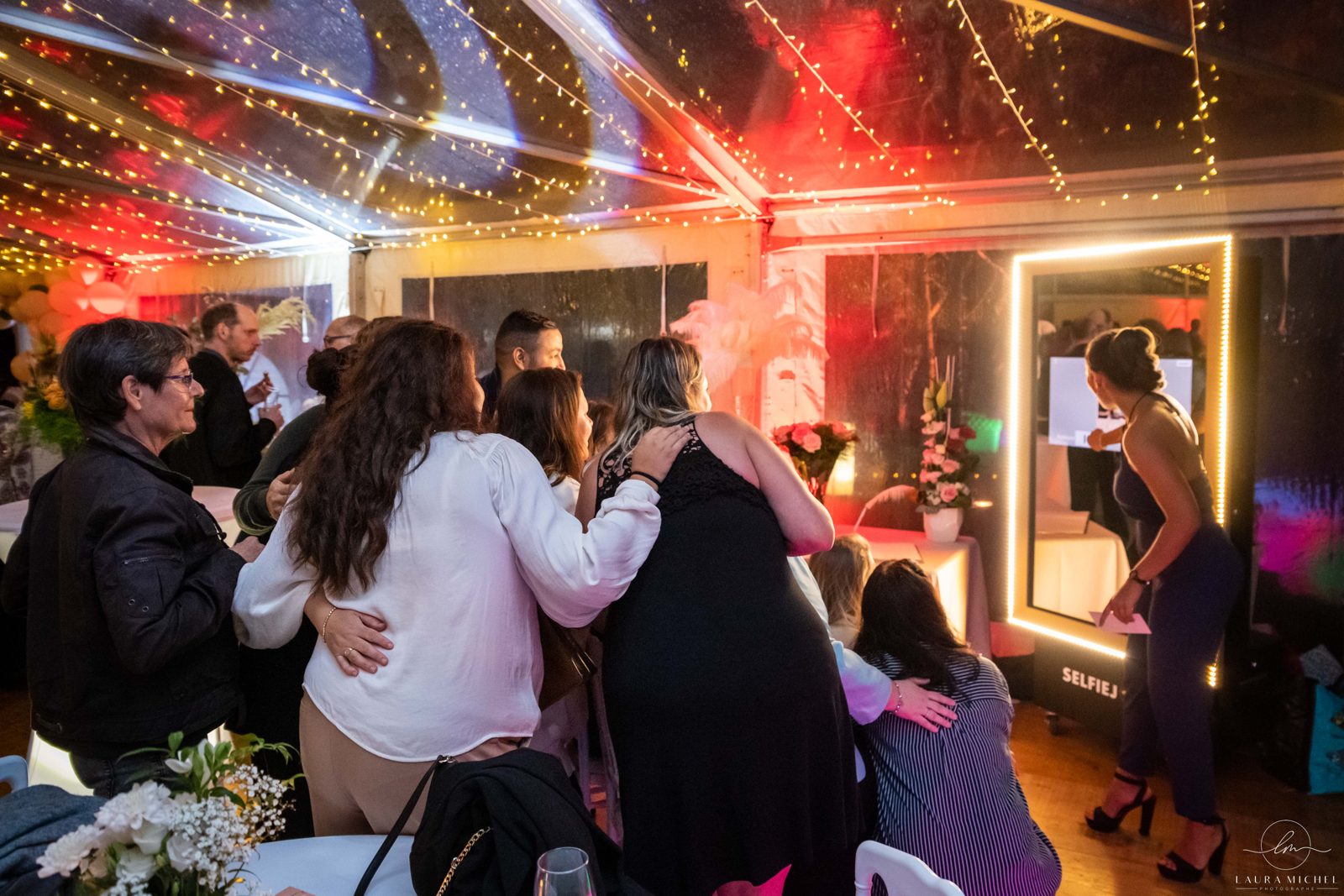 Invités devant une borne photo miroir encadrée de lumières, sous un chapiteau transparent tendu de guirlandes lumineuses