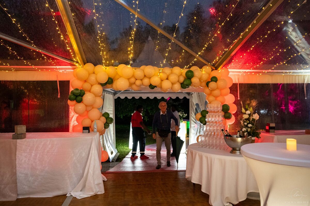 Entrée de chapiteau sous une arche de ballons pêche et verts, guirlandes lumineuses, mange-debout housé de blanc