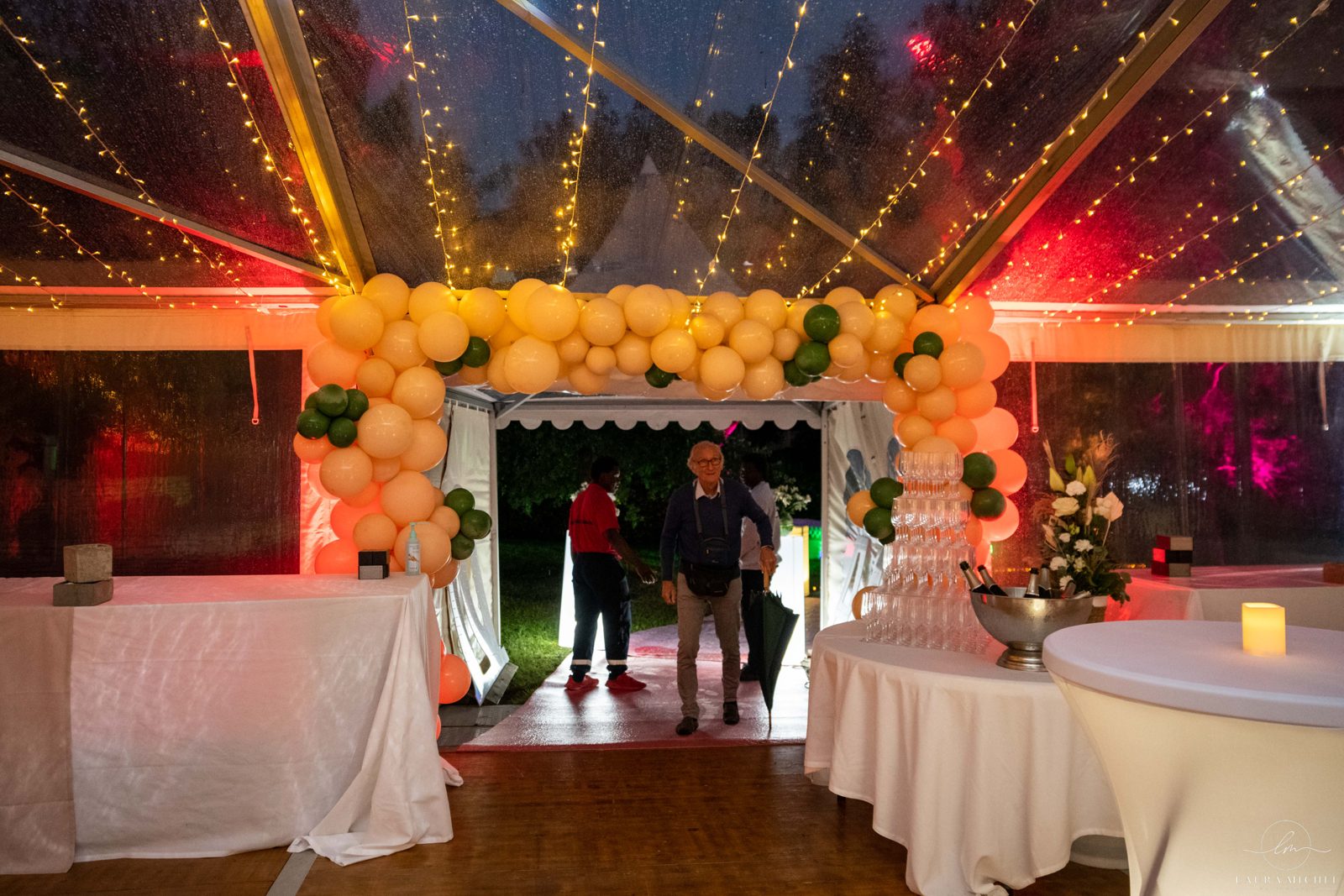 Entrée de chapiteau sous une arche de ballons pêche et verts, guirlandes lumineuses, mange-debout housé de blanc