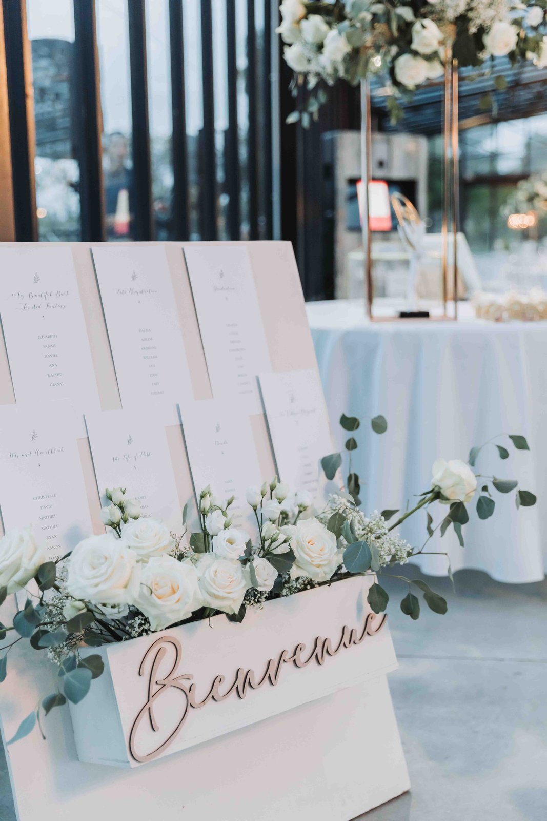 Panneau de bienvenue blanc garni de roses blanches et d’eucalyptus, plan de table en cartes calligraphiées au-dessus
