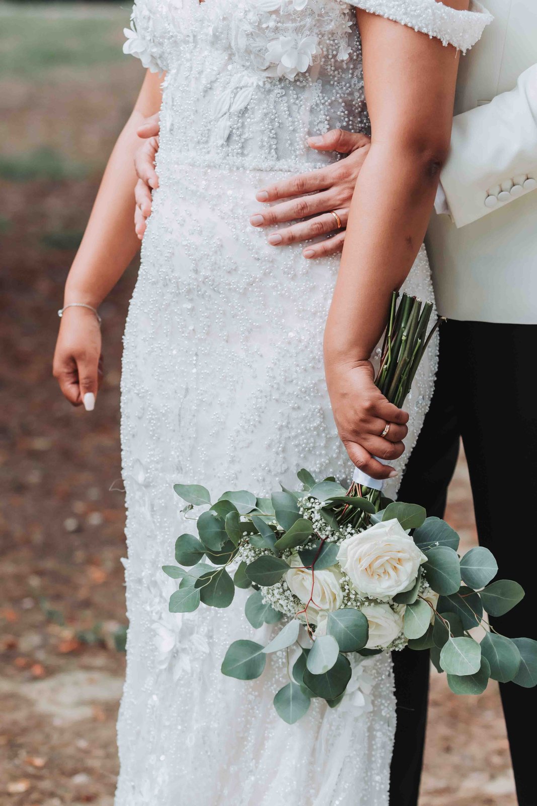 Bouquet de mariée de roses blanches et d’eucalyptus tenu contre une robe brodée, en plein air